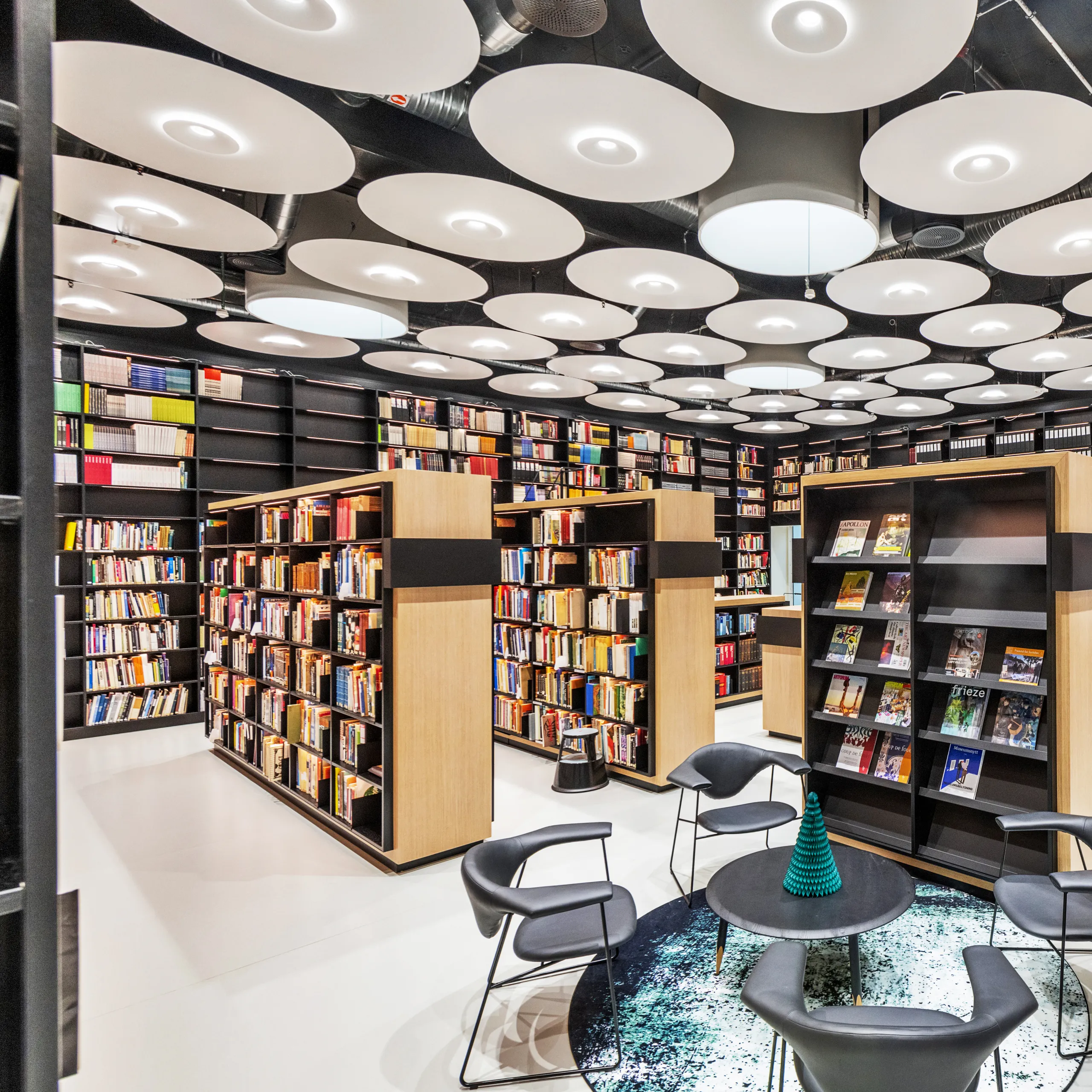 A vibrant library interior featuring numerous circular white acoustic ceiling islands arranged across a dark soffit, with timber bookcases and lounge seating below.