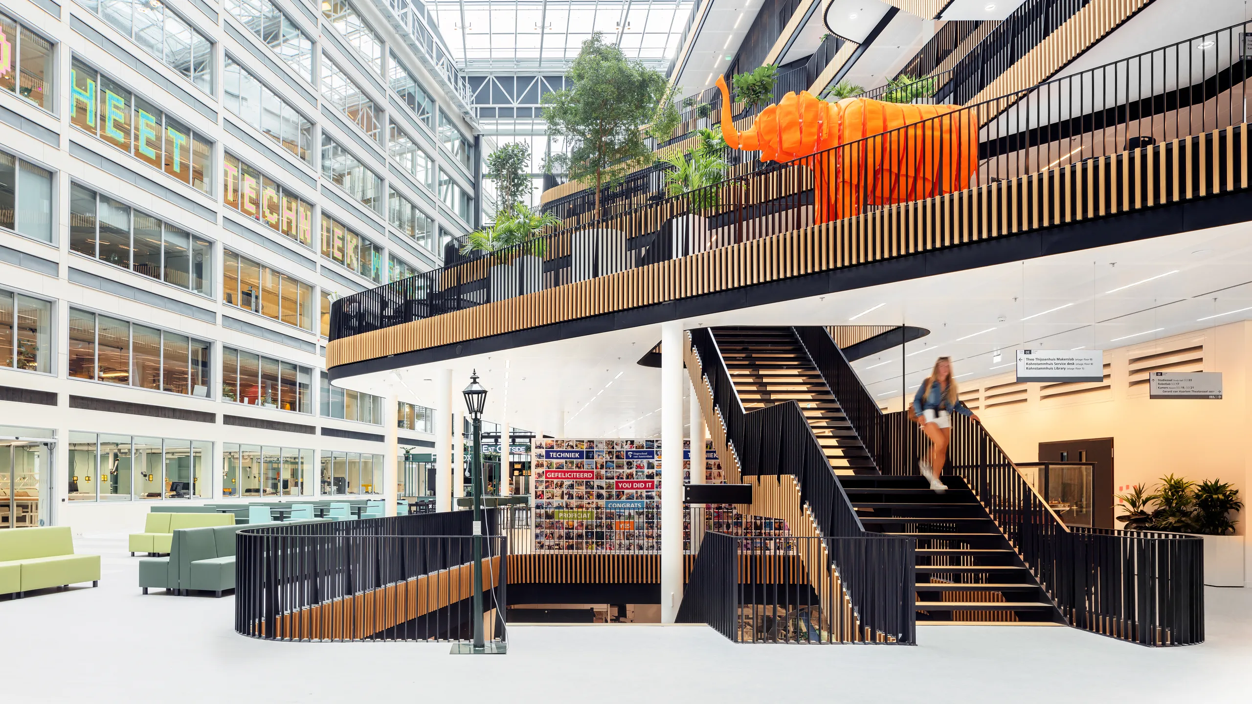 Large office atrium with a glass roof, central staircase, tiered walkways with planting, and an open, multi-level workspace filled with natural daylight.