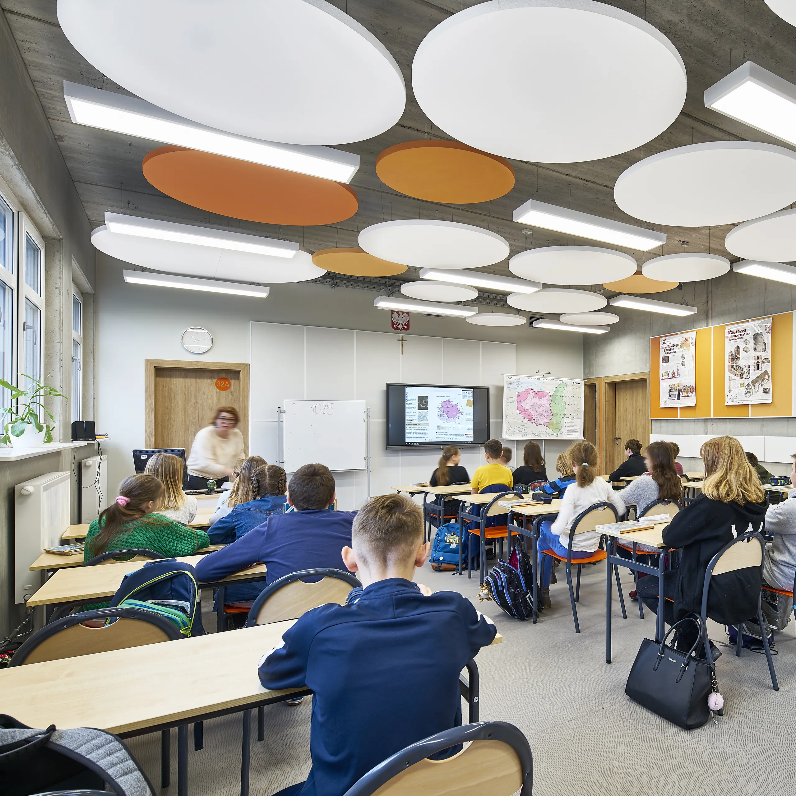 A modern classroom featuring circular acoustic ceiling islands in white and orange, suspended from an exposed soffit, with students seated at desks facing an interactive screen.