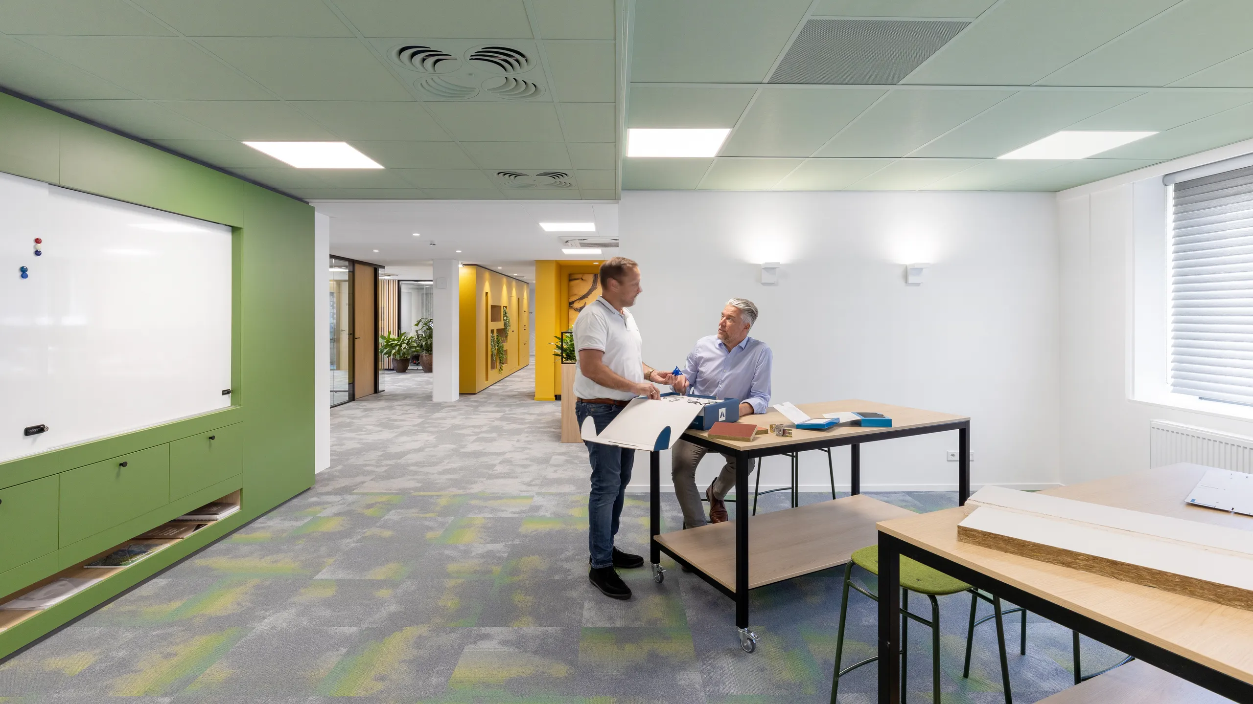 Two people collaborating at a standing desk in a modern office with green accents, whiteboard, and glass partitions.