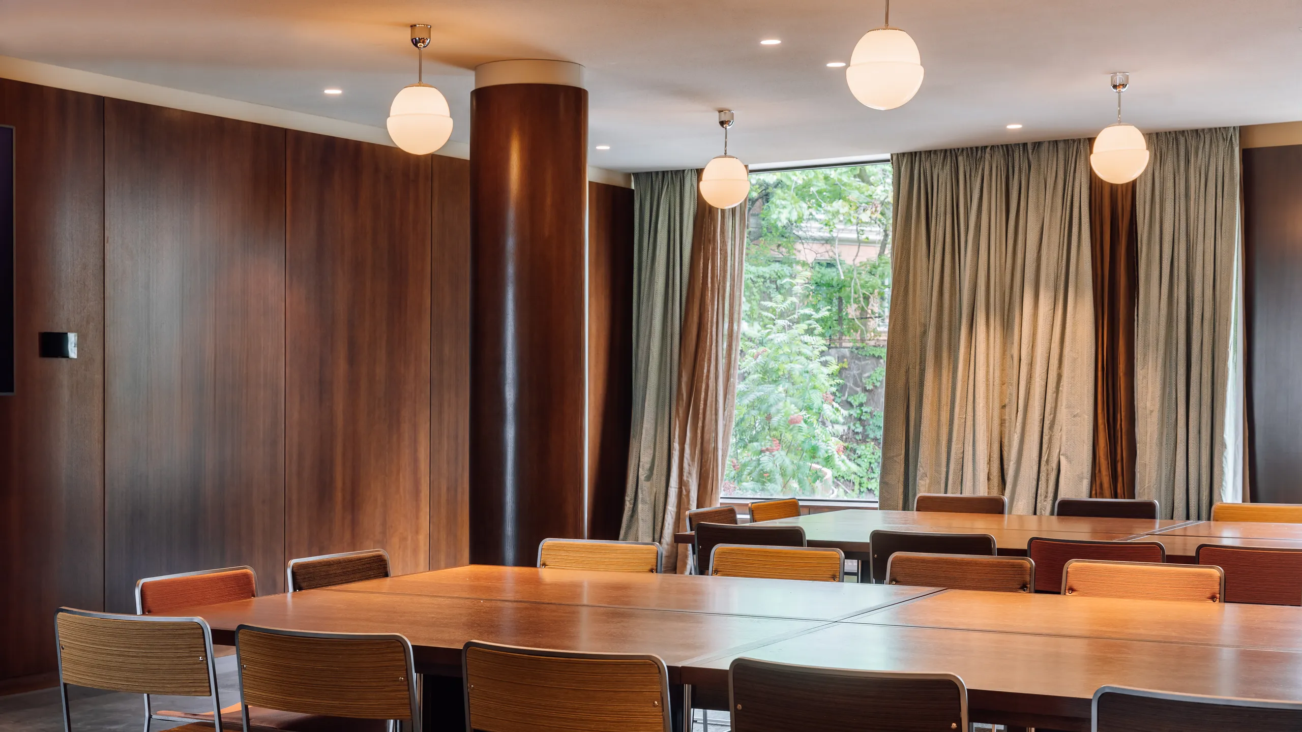 Meeting room with a smooth monolithic acoustic ceiling, wooden tables and chairs, pendant lighting, and large windows with curtains overlooking greenery.