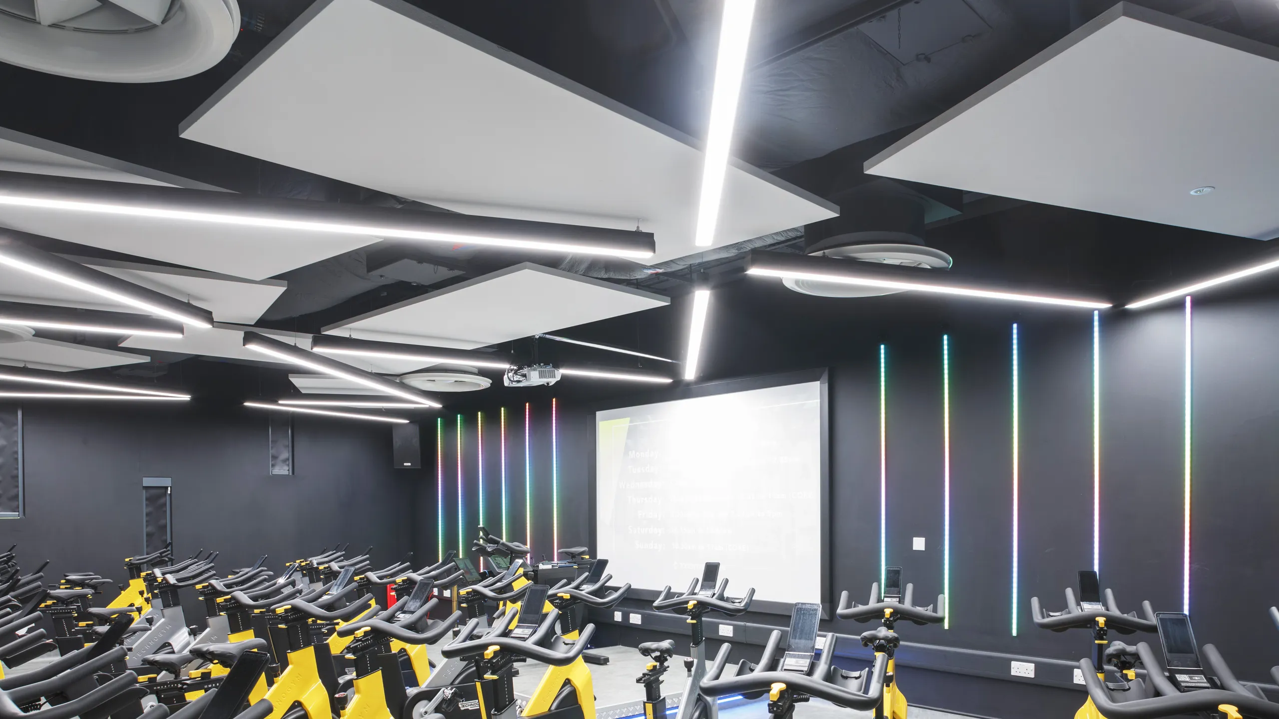 Modern indoor cycling studio with white floating ceiling panels, rows of stationary bikes, vibrant LED light strips, and geometric ceiling panels.