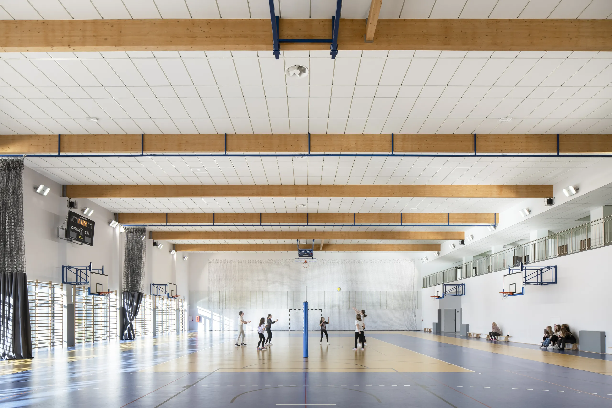 A spacious sports hall featuring acoustic ceiling tiles fitted between large timber beams, with natural light, blue-and-yellow court markings, and students playing volleyball.