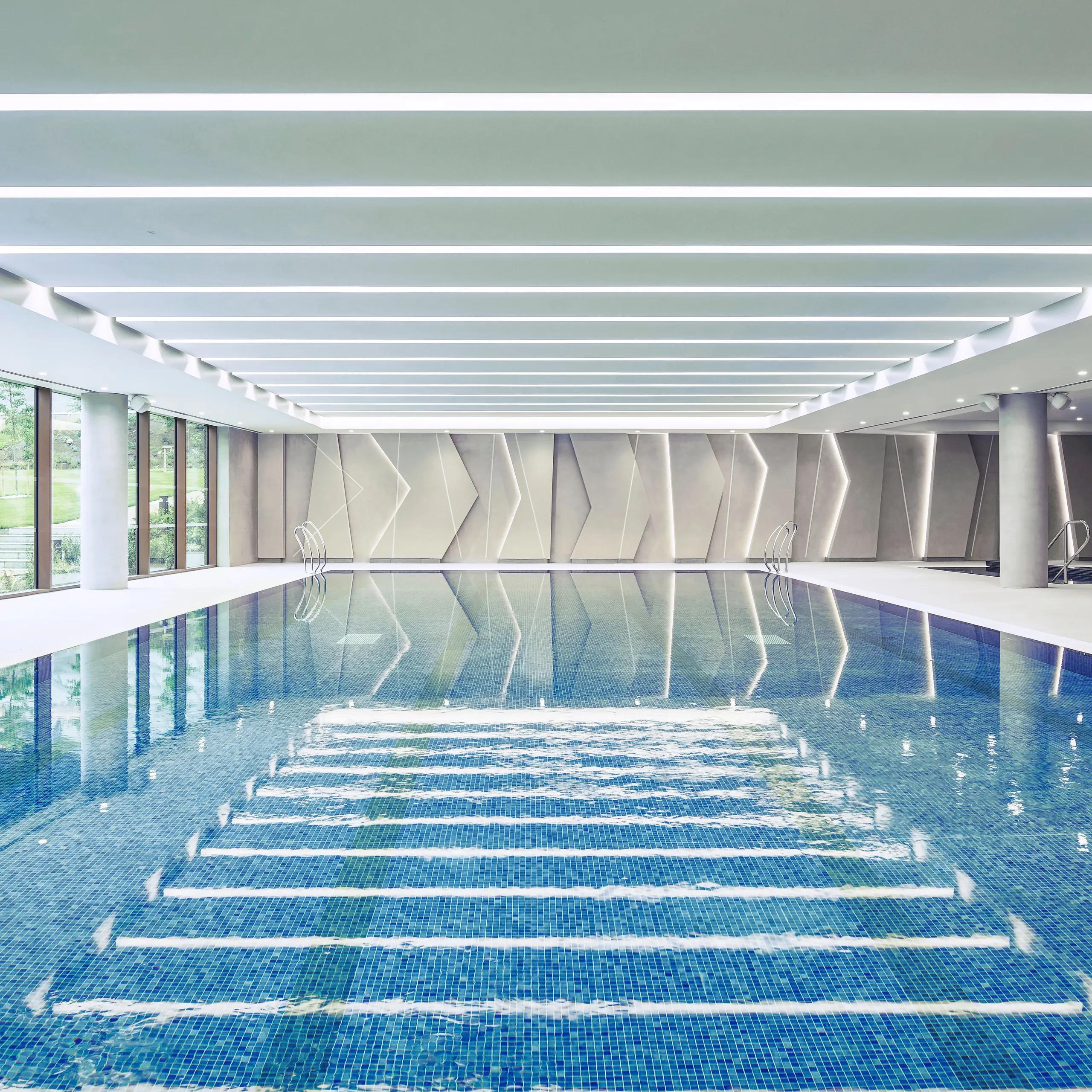 Indoor swimming pool with a long lap pool, large side windows, geometric wall panels, and a white linear acoustic ceiling with integrated lighting reflected in the water.