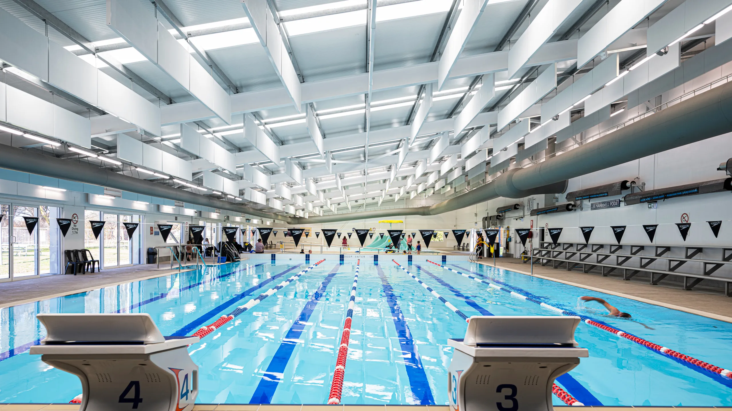 Indoor swimming pool with suspended white acoustic baffles across the ceiling, lane markings, starting blocks, and spectator seating along the poolside.