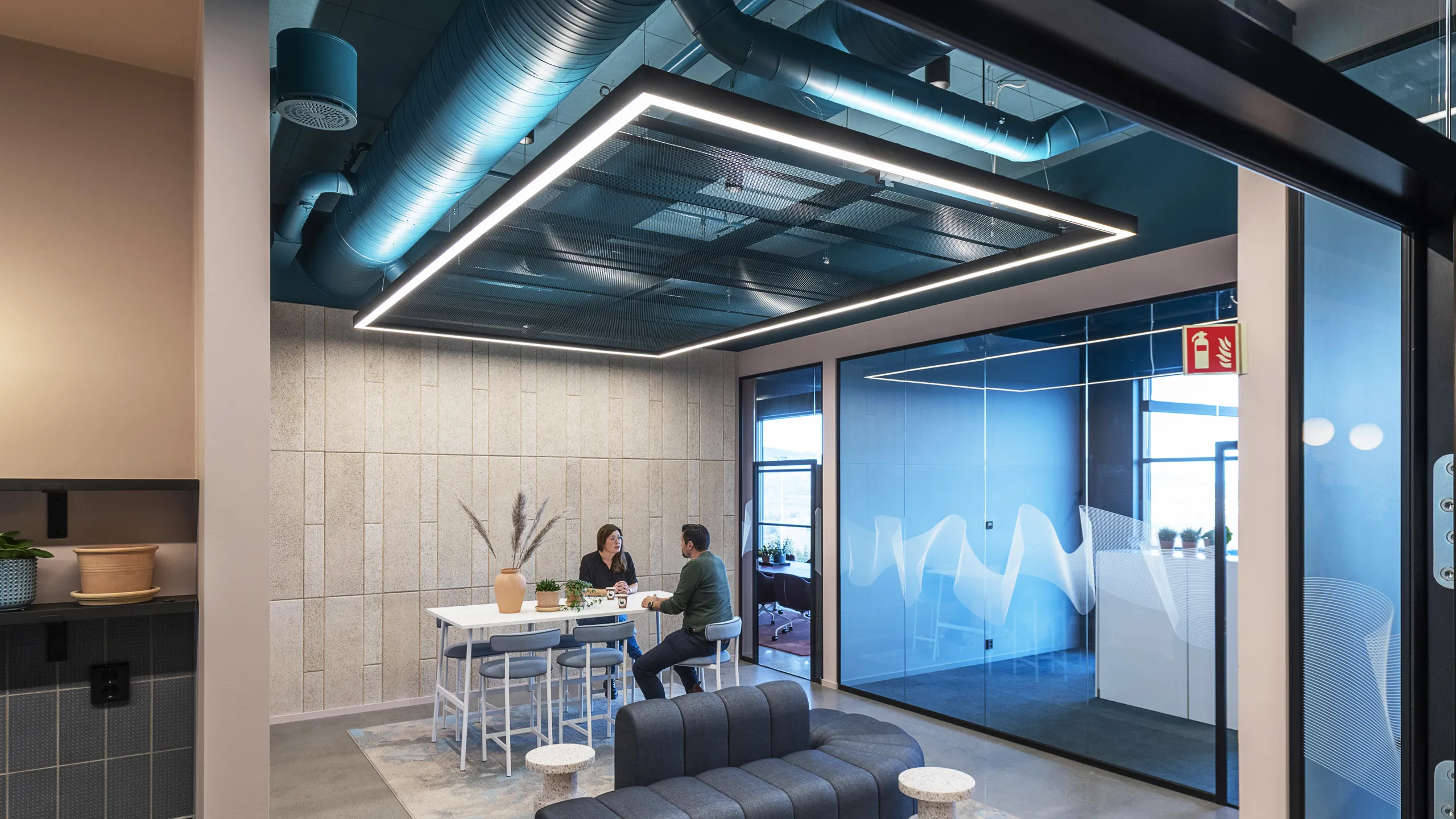 Modern office lounge with two people sitting at a high table, an exposed open plenum blue office ceiling, blue accents, a cushioned bench, and large windows.