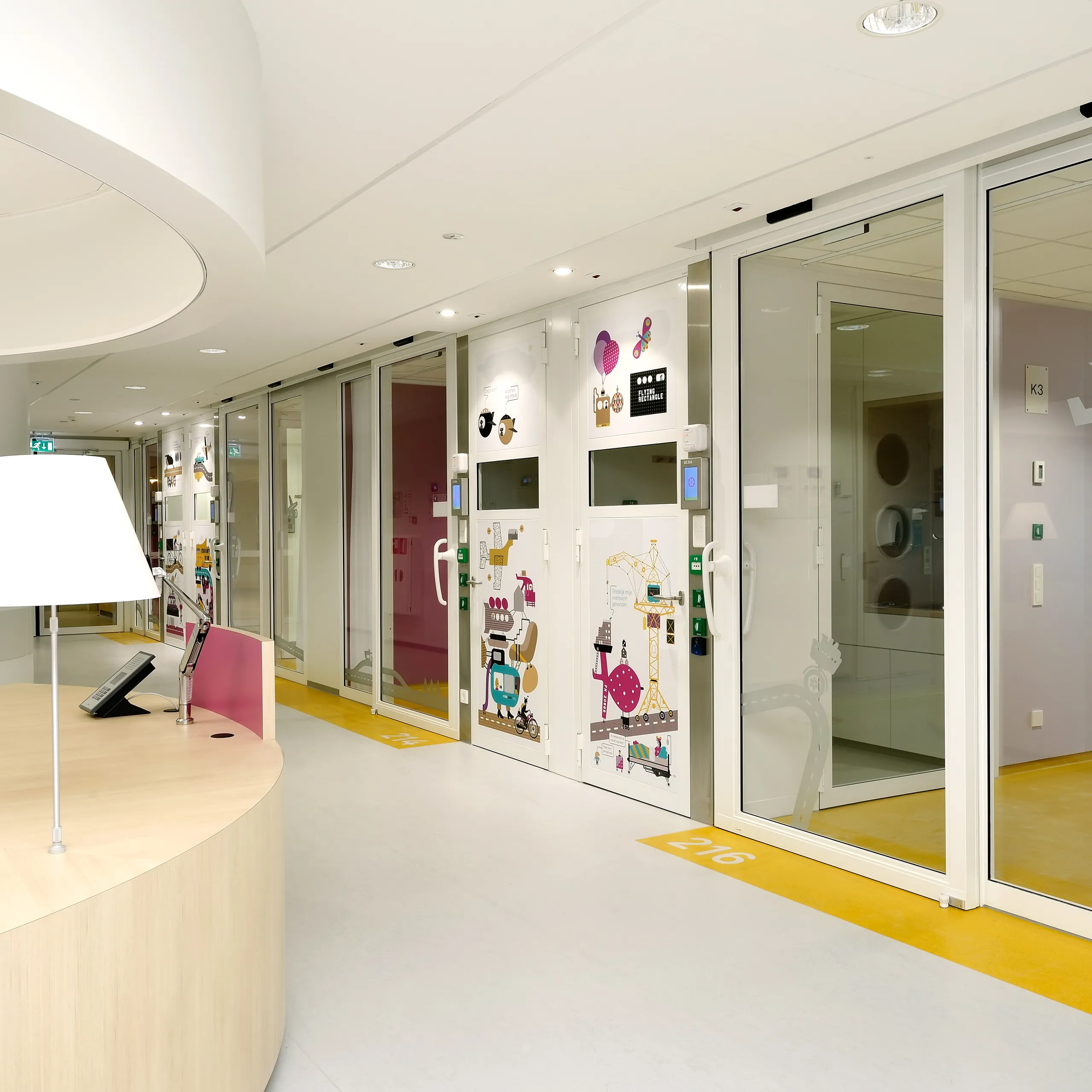 Children’s hospital corridor with a curved wooden nurse desk, bright white acoustic ceiling panels, and glass room doors decorated with colourful illustrations and icons.