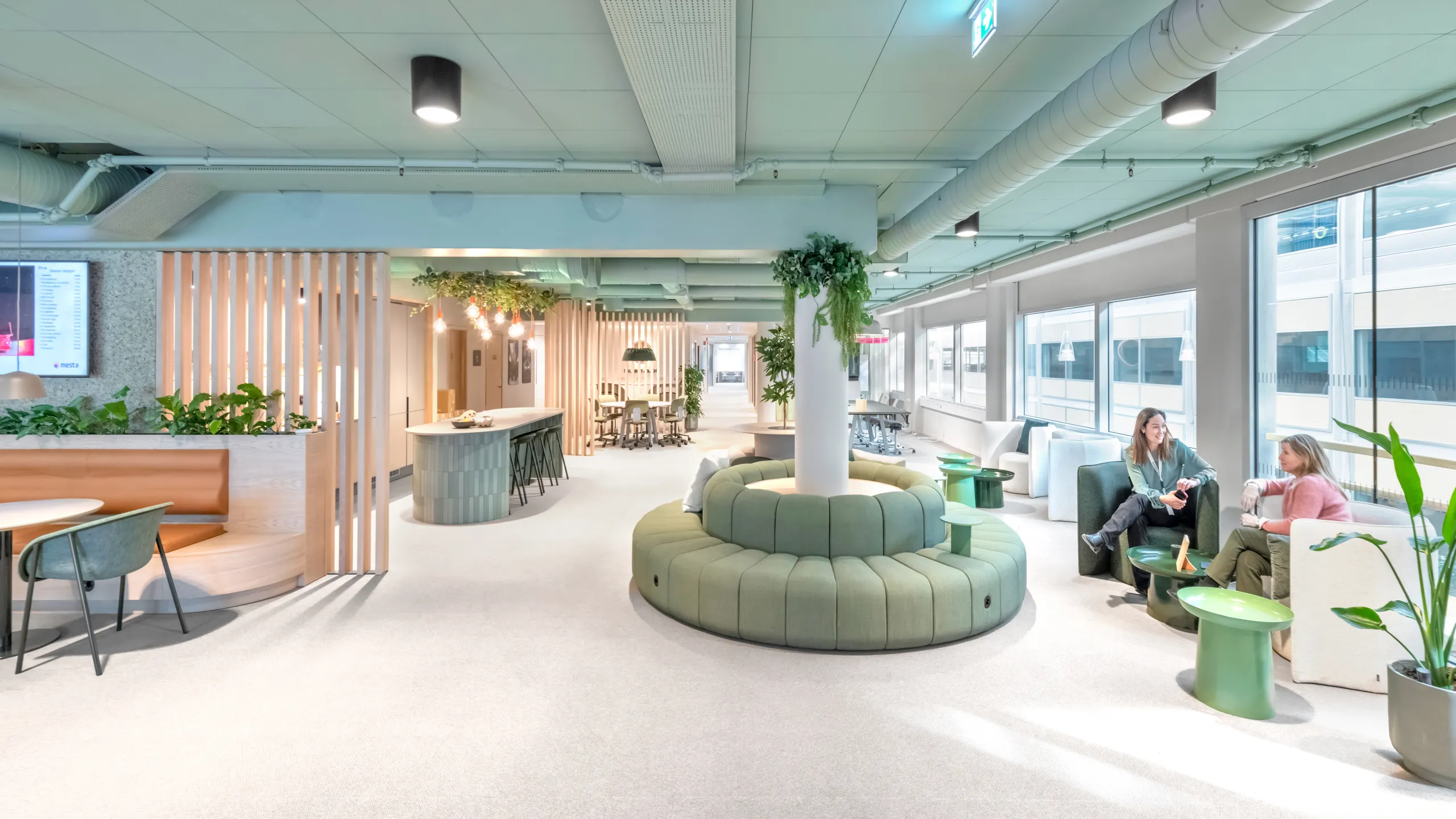 Modern open-plan office with pastel green ceiling, green seating, plants, and large windows. Two people are seated and conversing on a sofa to the right.