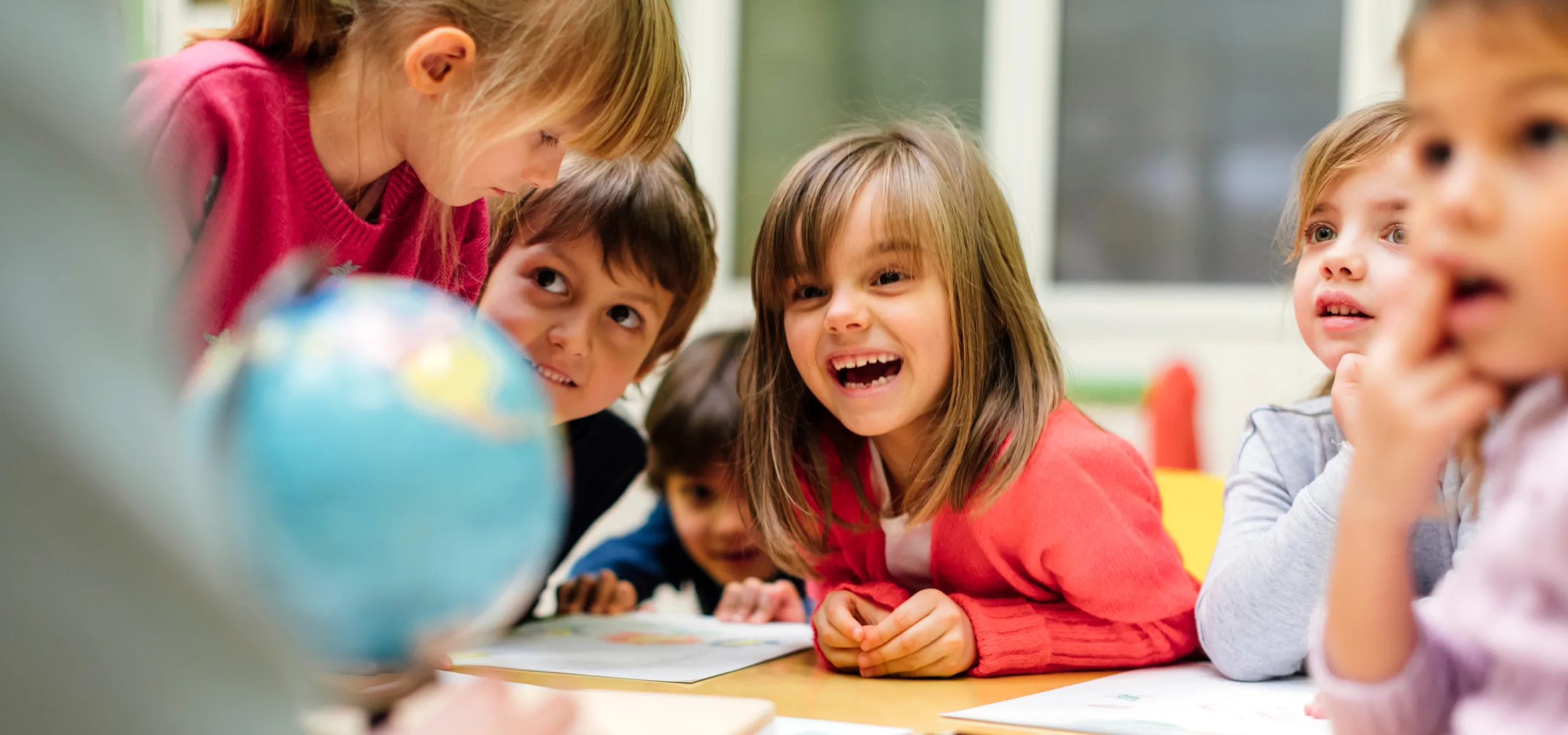 A group of young children sit around a classroom table, smiling and leaning in with curiosity as they look at a small globe and work on drawings.