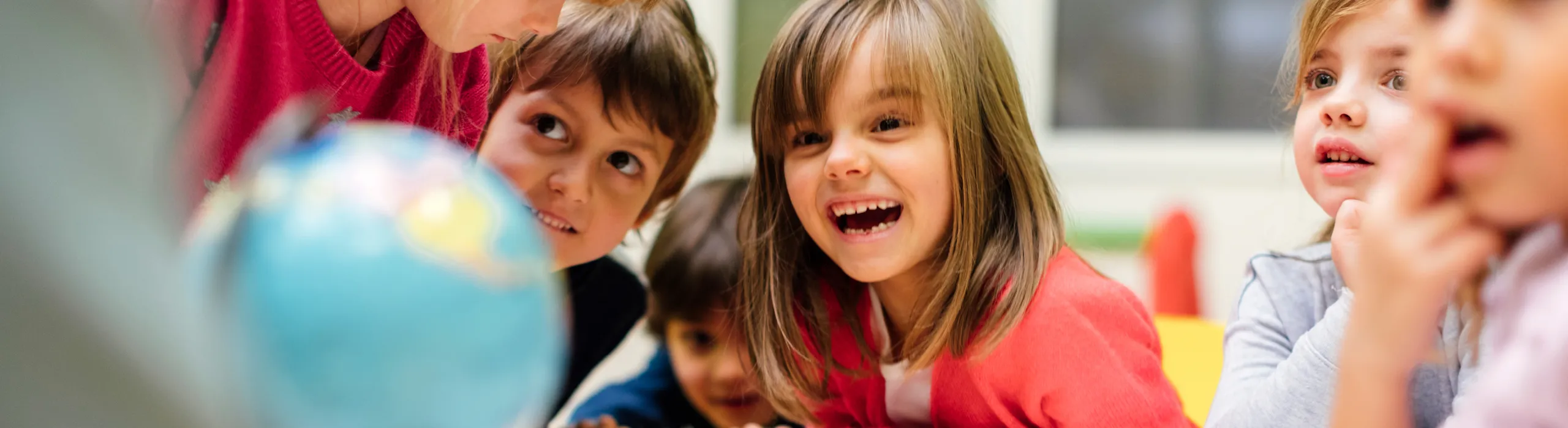 A group of young children sit around a classroom table, smiling and leaning in with curiosity as they look at a small globe and work on drawings.