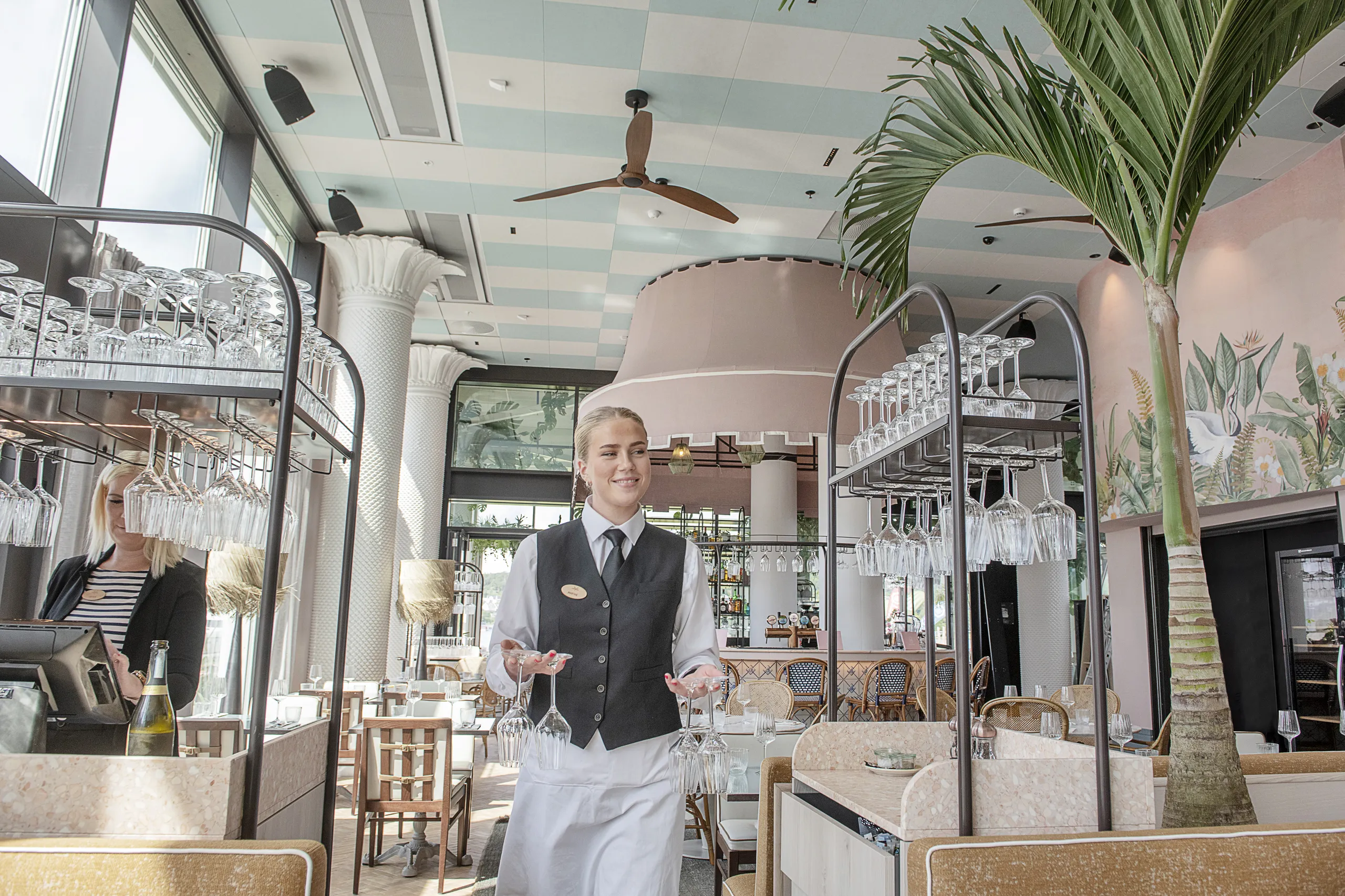 Restaurant interior with a patterned acoustic ceiling and ceiling fans above a bright dining area, where a server carries wine glasses between tables and bar carts.