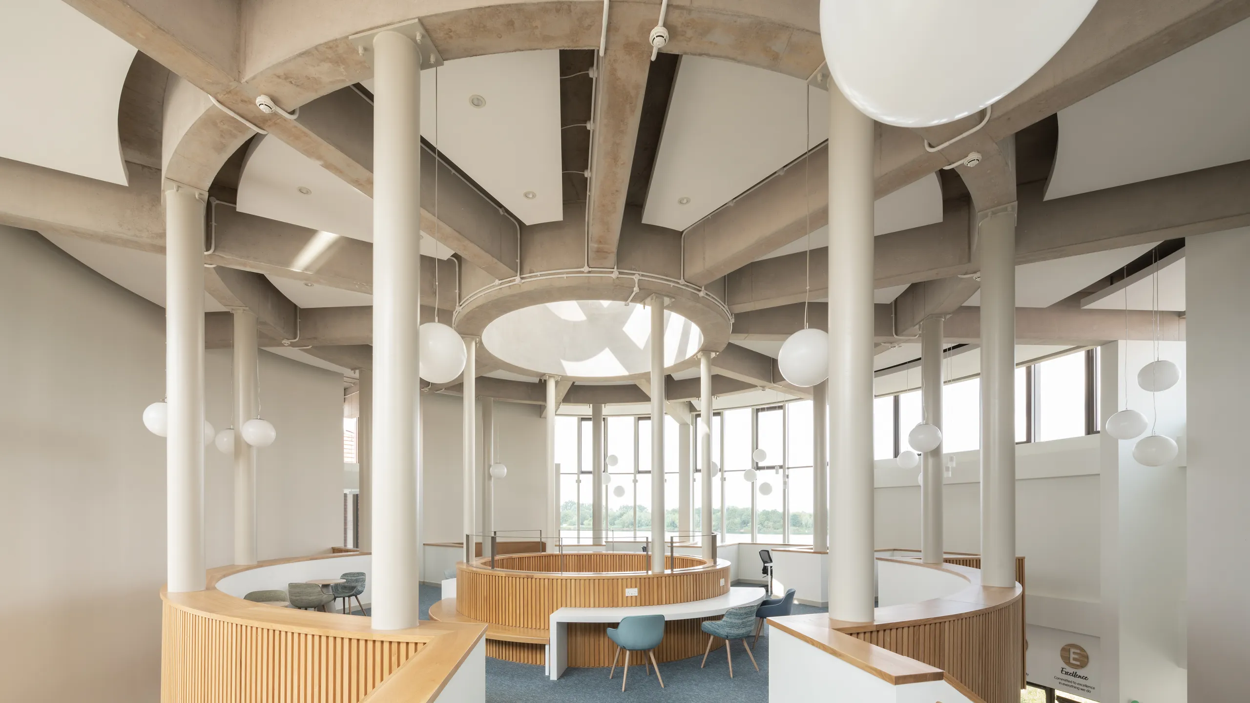 Spacious modern interior with white floating ceiling panels, circular wooden desks, white pillars, large windows, and hanging globe lights. Blue carpet and light gray walls.