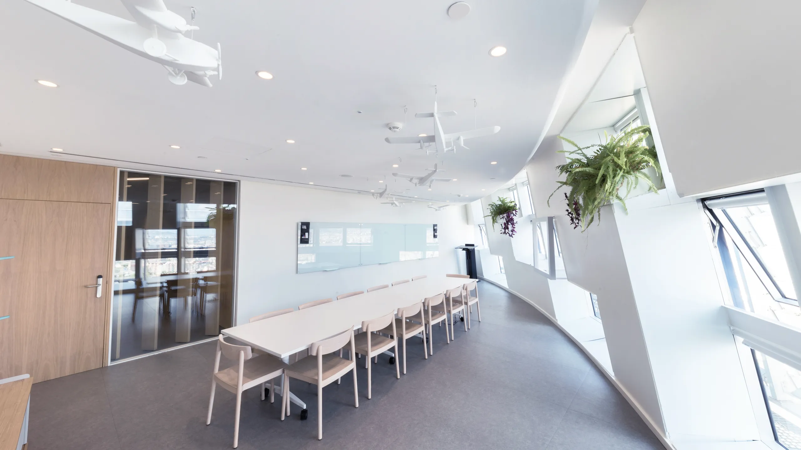 Meeting room with a curved monolithic acoustic ceiling, long table with chairs, suspended model planes, and angled windows bringing in natural daylight.