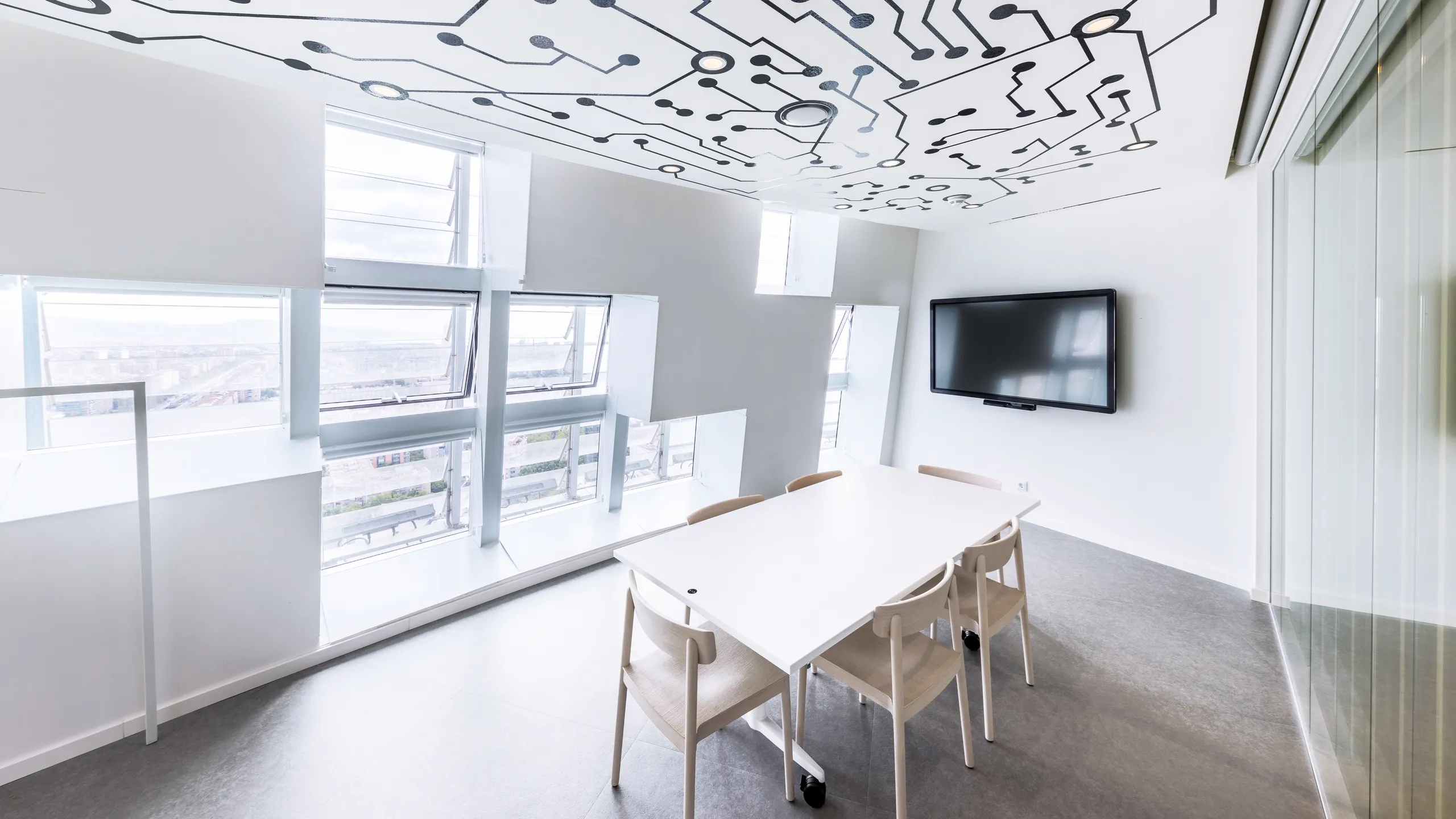 Meeting room with acoustic ceiling tiles featuring a printed circuit pattern, table and chairs, wall-mounted screen, and angled windows bringing in natural daylight.