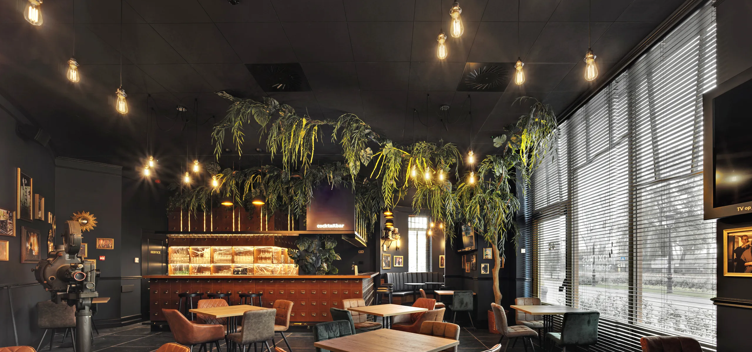 Cozy café interior with black suspended ceiling, modern decor, wooden tables, brown chairs, hanging plants, and warm lighting from exposed bulbs. Large windows on the right.