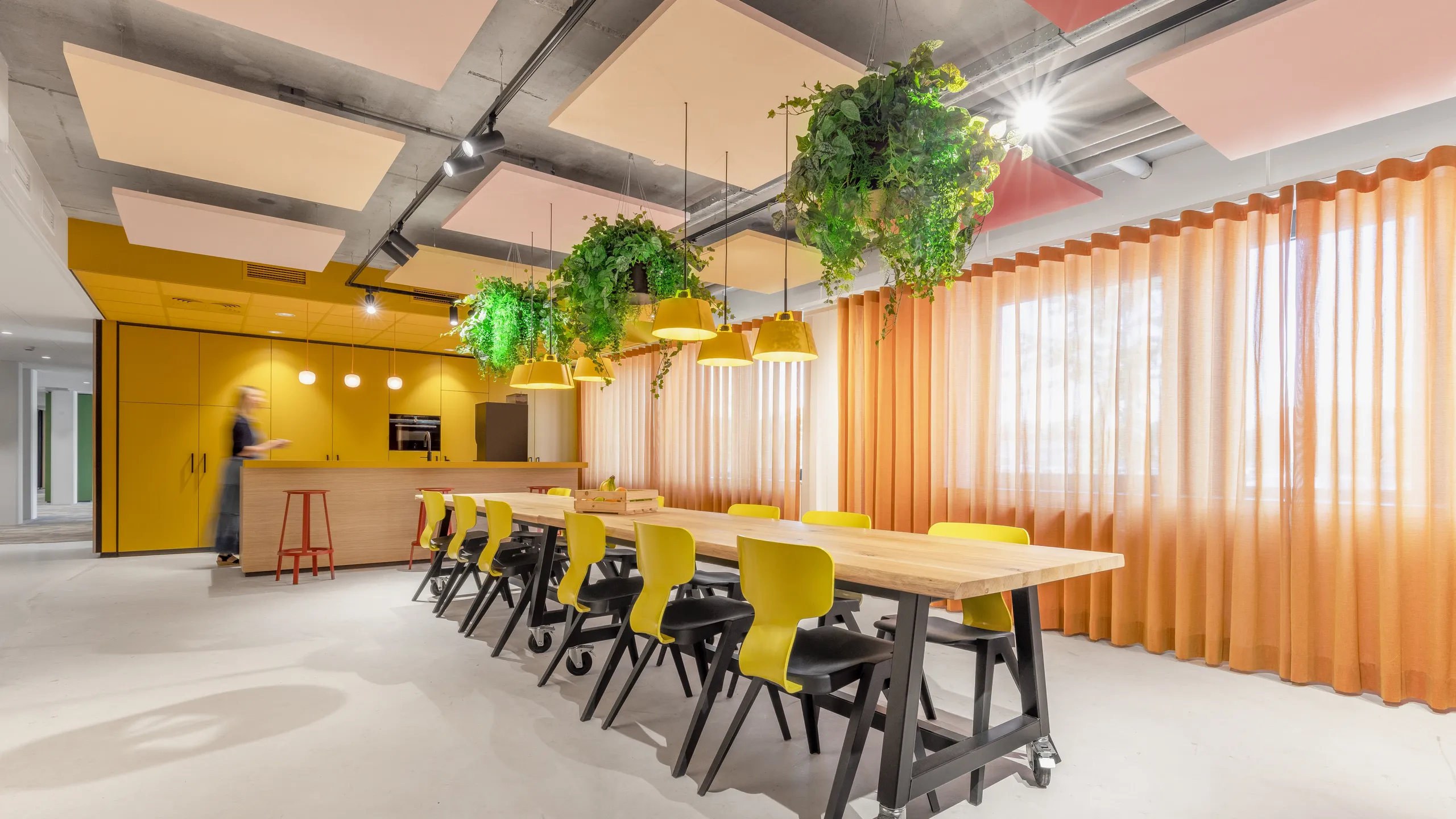Modern office kitchen with a long wooden table, pink floating ceiling panels, yellow chairs, overhead plants, yellow cabinets, and large windows with sheer curtains.