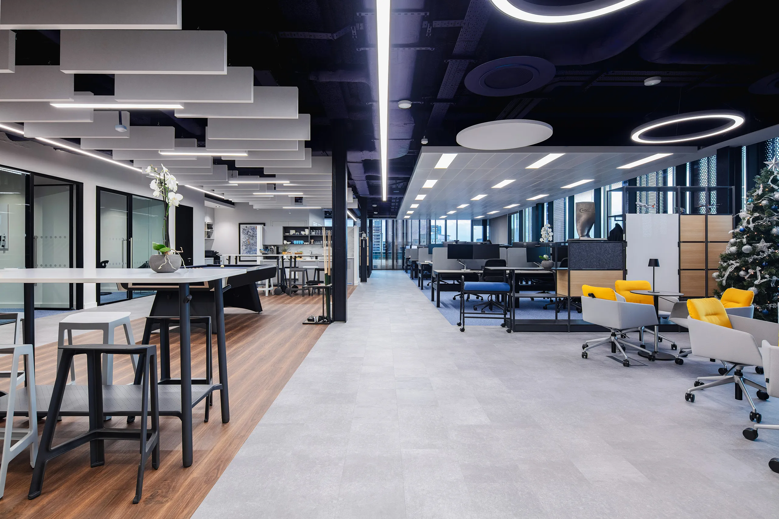 Modern open office with ceiling-mounted acoustic baffles and round acoustic islands, combining lounge seating, shared tables, and linear lighting under a dark exposed ceiling