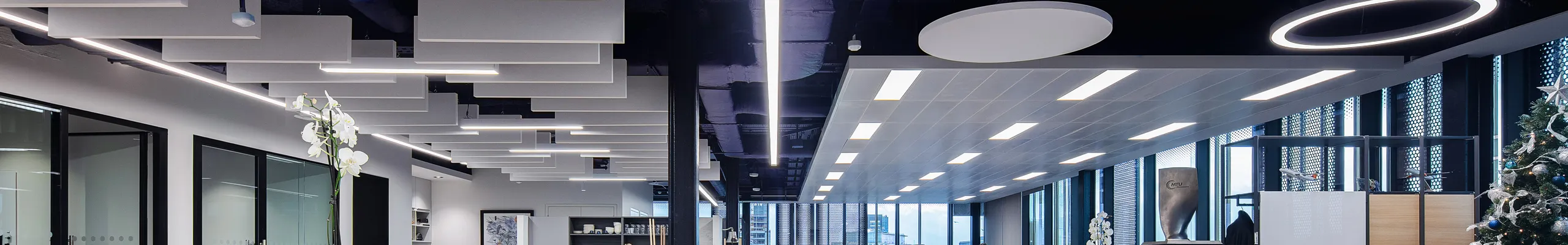 Modern open office with ceiling-mounted acoustic baffles and round acoustic islands, combining lounge seating, shared tables, and linear lighting under a dark exposed ceiling