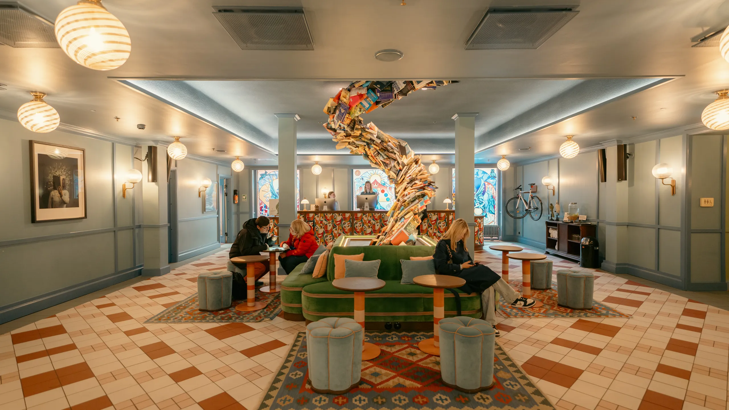 Hotel lobby lounge with a smooth monolithic acoustic ceiling, patterned tile floor, green seating, and a sculptural installation made from stacked books at the centre of the space.