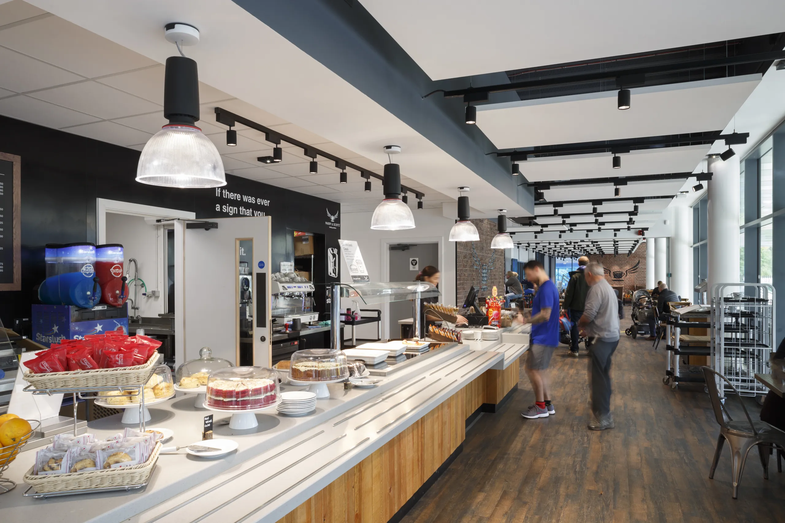 Cafeteria interior with a self-service food counter, seating area, and a white acoustic tile ceiling with suspended lighting above the space.