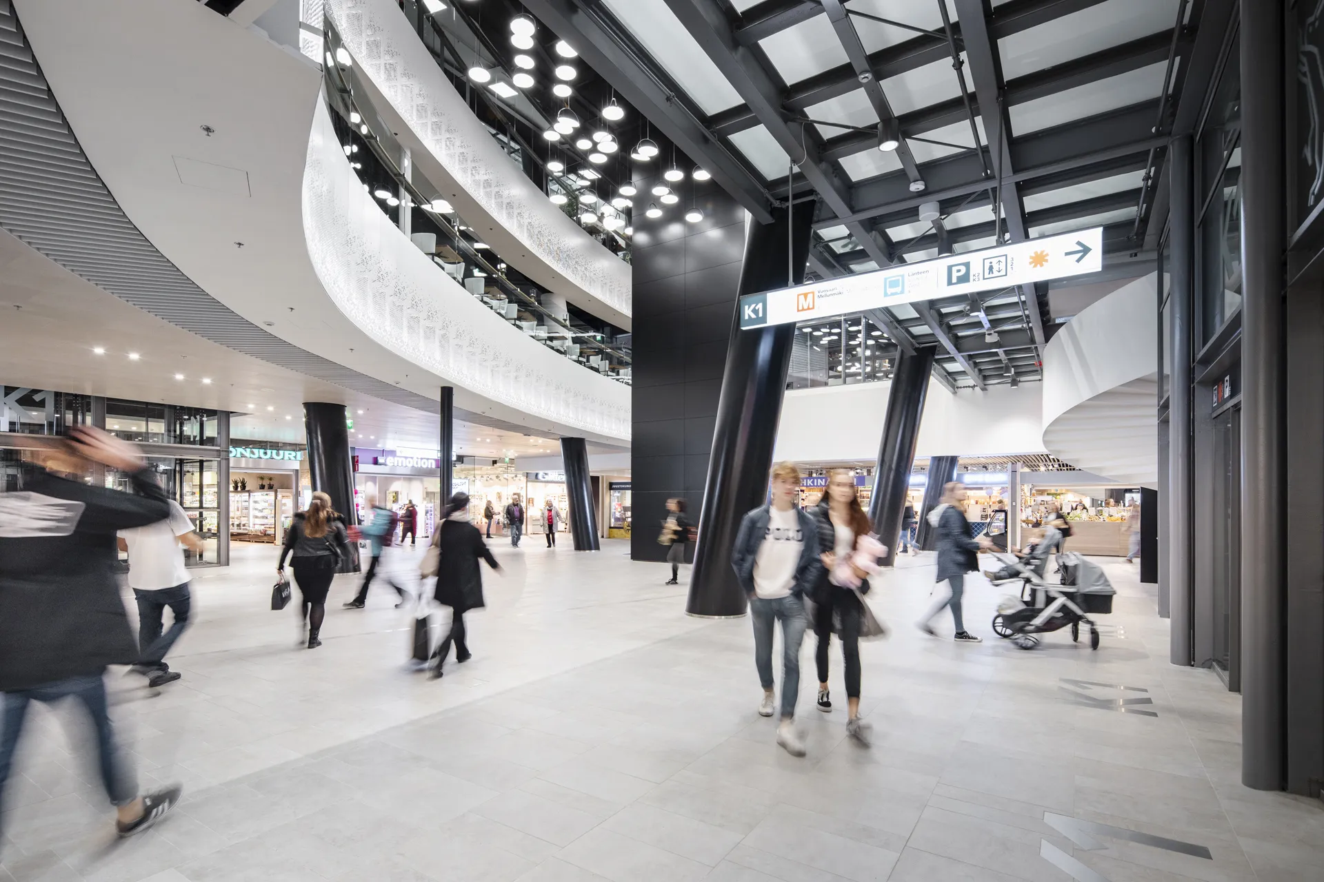 A bright shopping mall concourse featuring sweeping curved white acoustic ceilings and dramatic black steel columns, with shoppers moving between illuminated store entrances.
