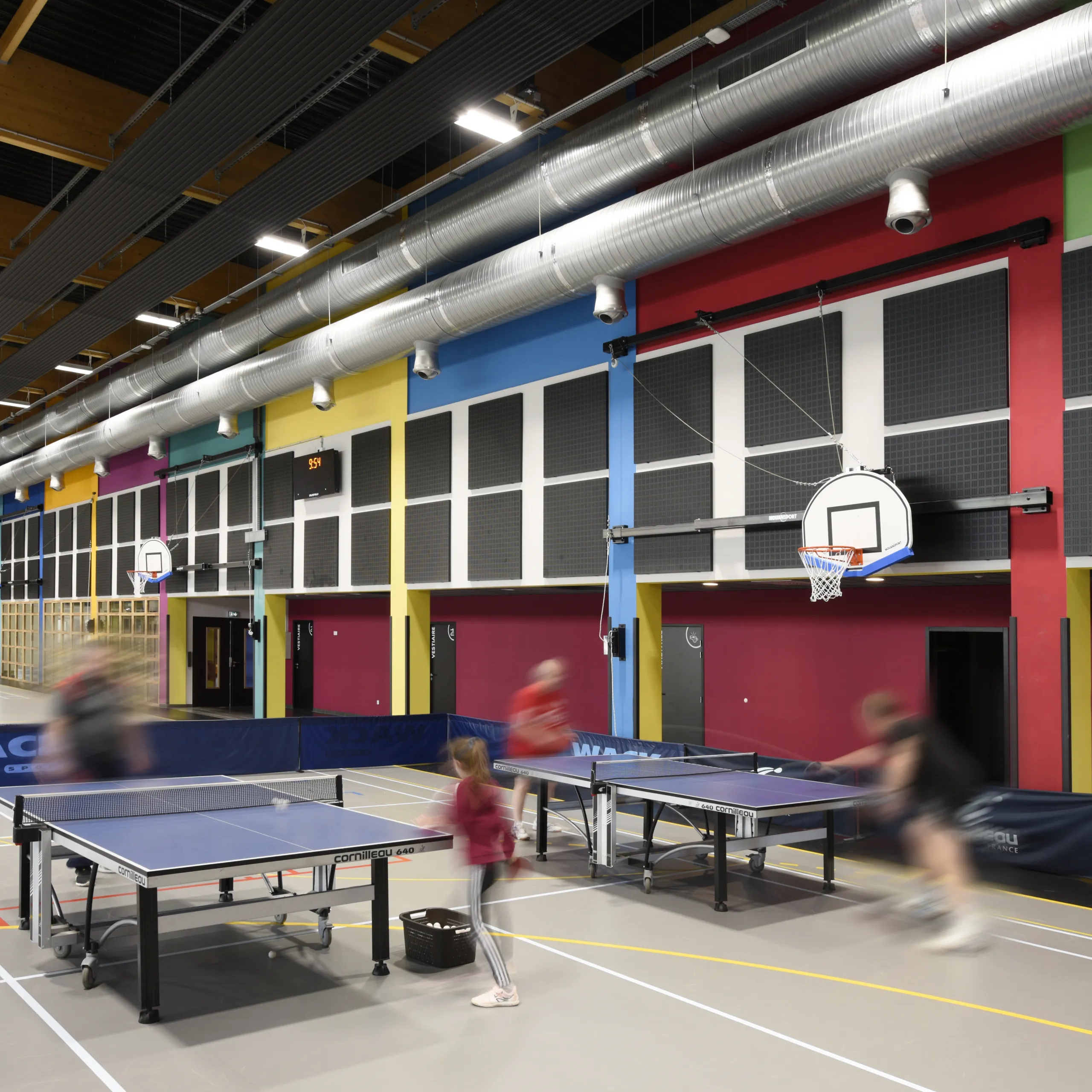 Multi-purpose sports hall with colourful wall panels, exposed ventilation ducts, and wall-mounted acoustic panels, where people play table tennis beneath basketball hoops.