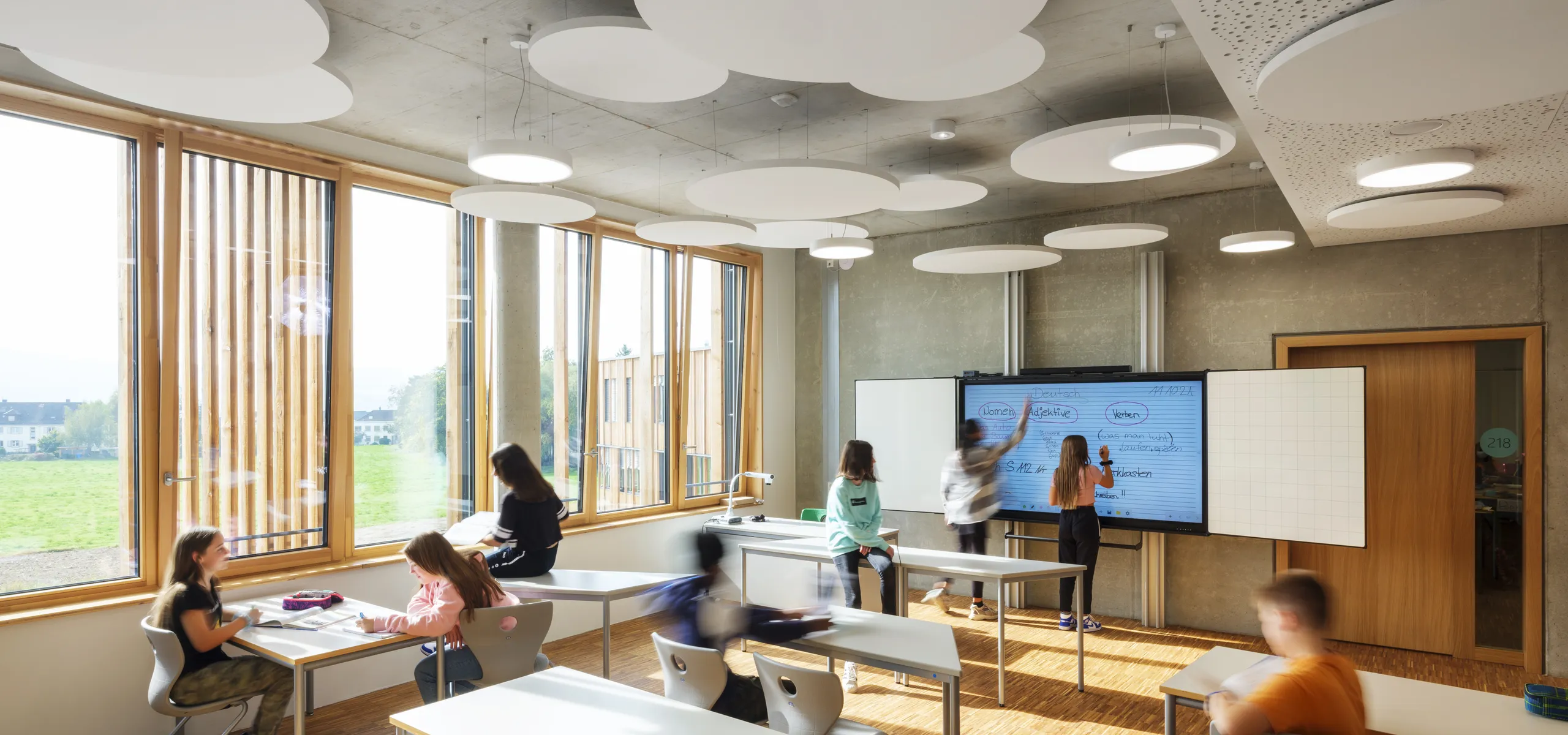 modern classroom featuring circular white acoustic ceiling islands suspended from an exposed concrete soffit, with large timber-framed windows and students working together at desks and a digital whiteboard