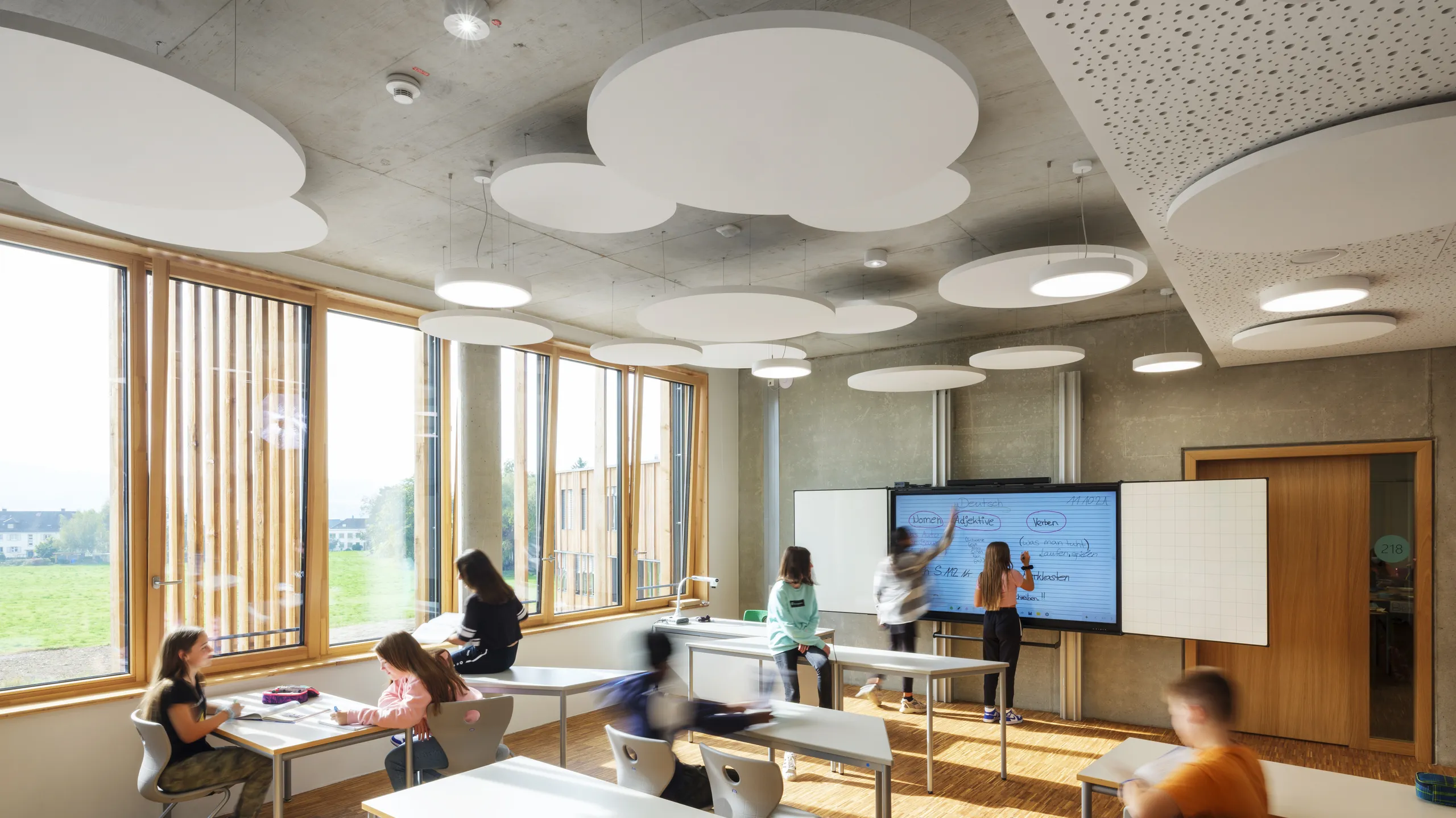 Modern classroom with floating ceiling panels, students engaged in various activities; features large windows, unique ceiling lights, and a digital screen.