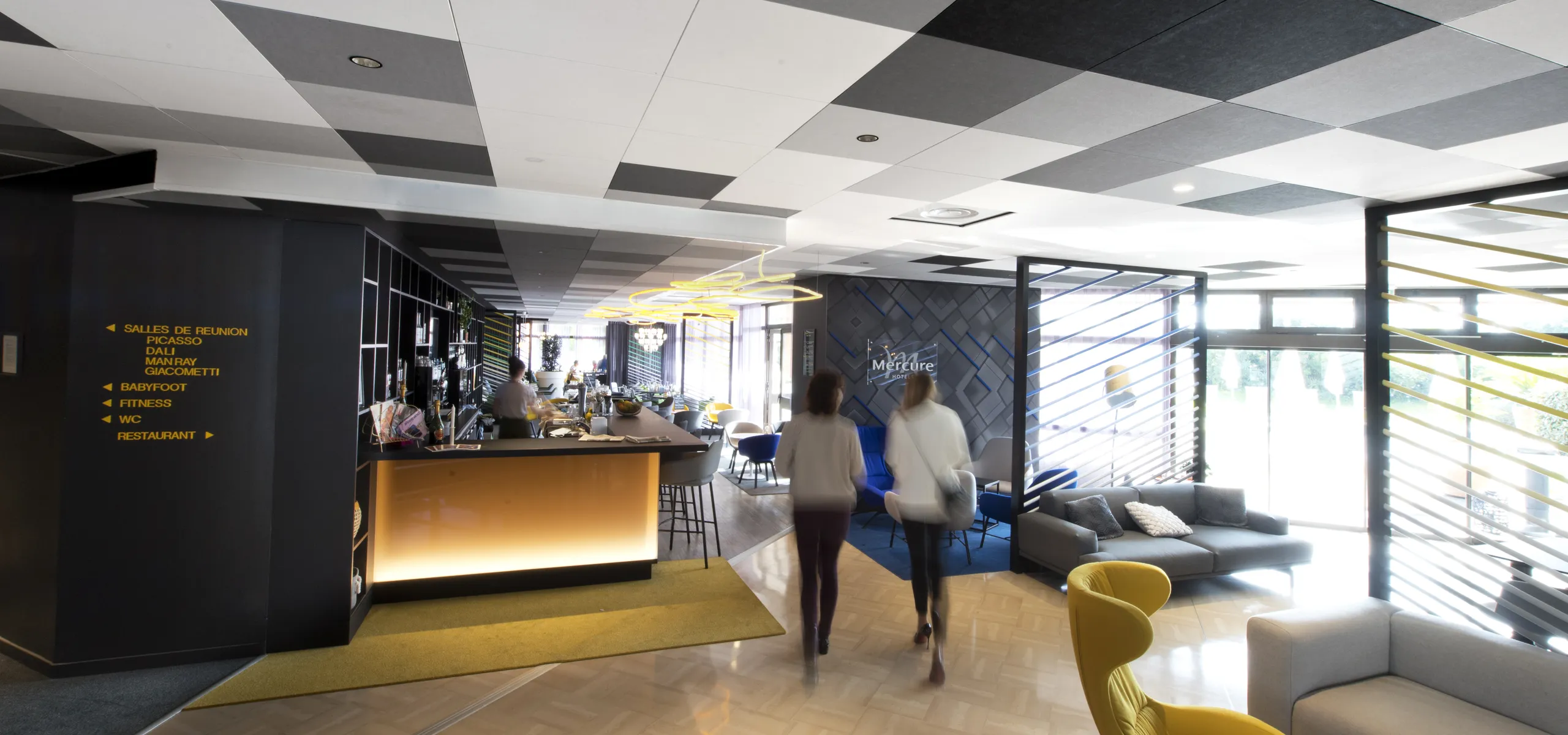Hotel bar and lounge with a black and white checkerboard acoustic ceiling, illuminated bar counter, seating areas, and large windows bringing daylight into the space.