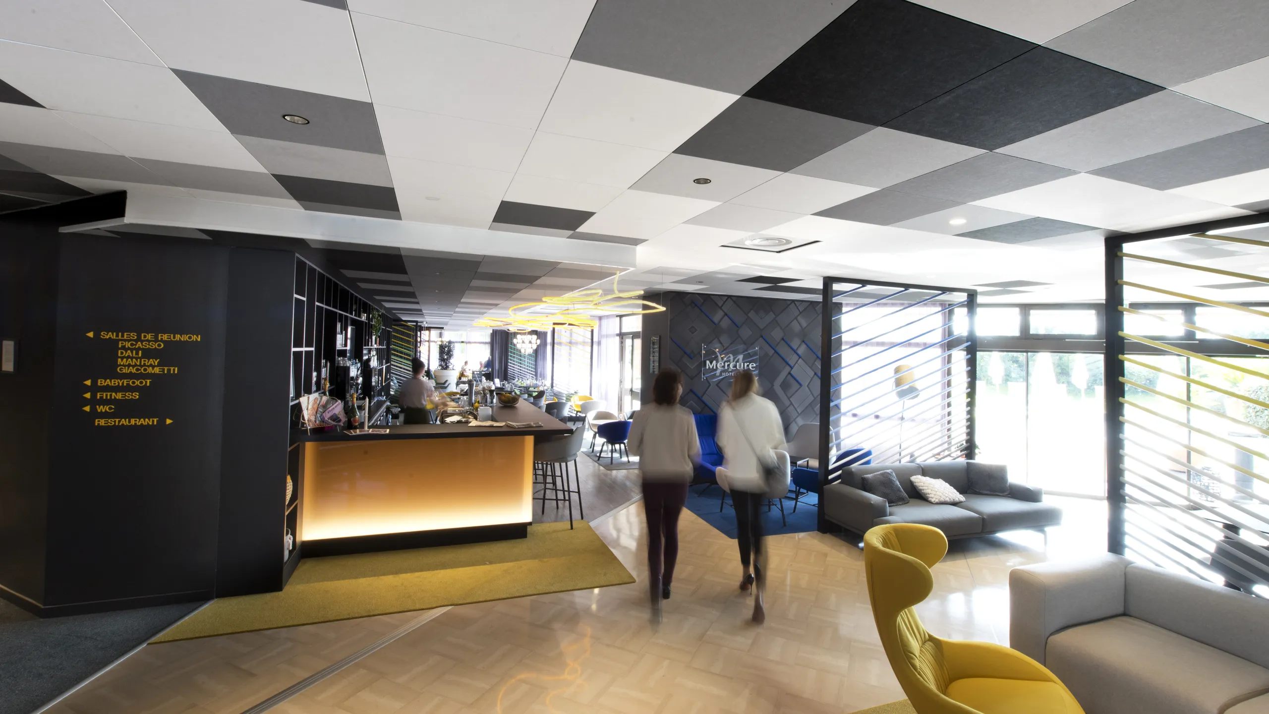 Hotel bar and lounge with a black and white checkerboard acoustic ceiling, illuminated bar counter, seating areas, and large windows bringing daylight into the space.