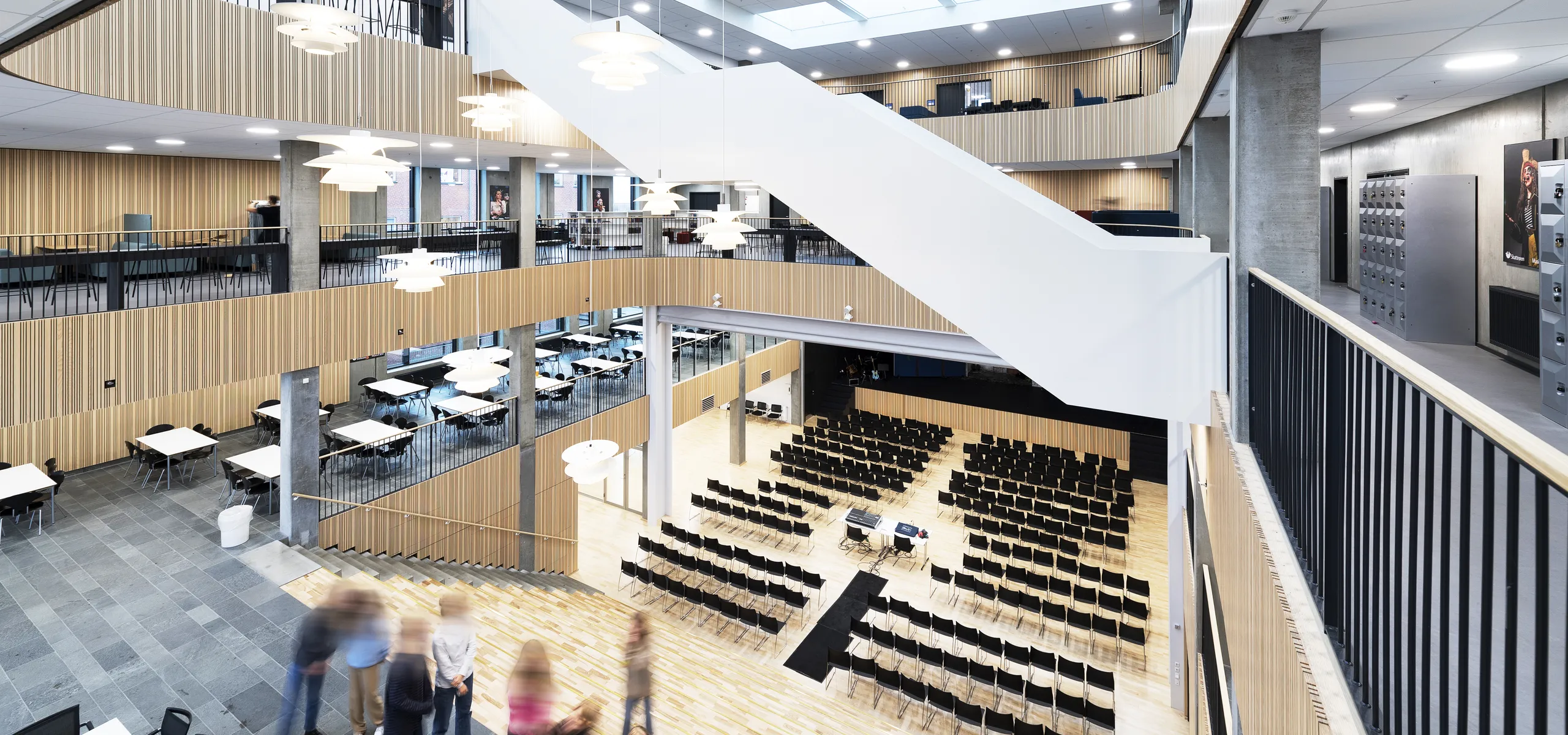 A spacious, multi-storey school atrium featuring white acoustic ceiling tiles and large skylights above open walkways, tiered seating, and a central hall arranged for an assembly