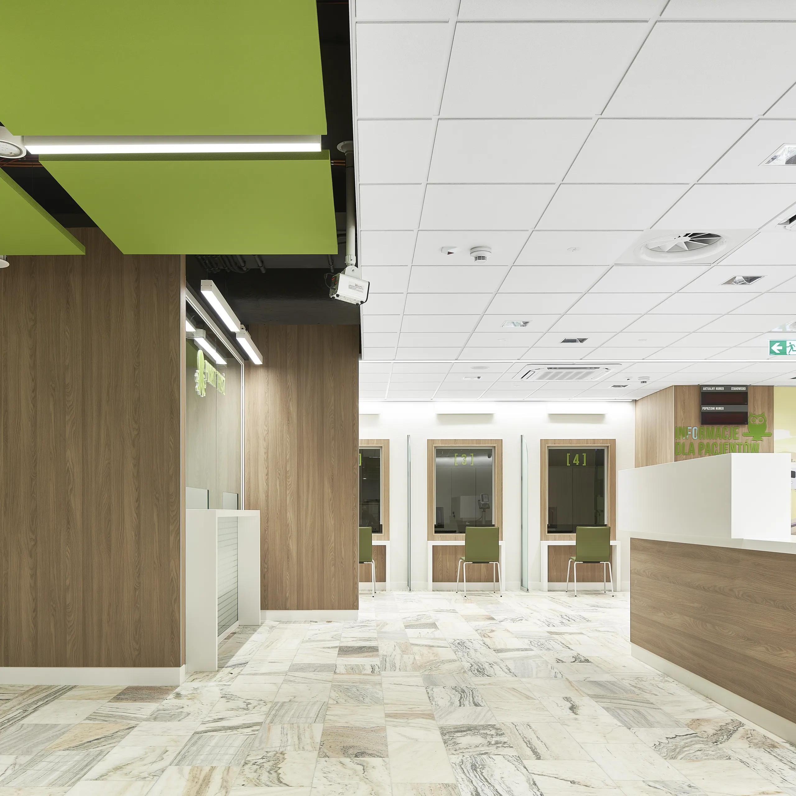 Hospital reception area featuring a white acoustic ceiling with integrated services and suspended green acoustic islands, complemented by a wood-panelled desk and green waiting benches.