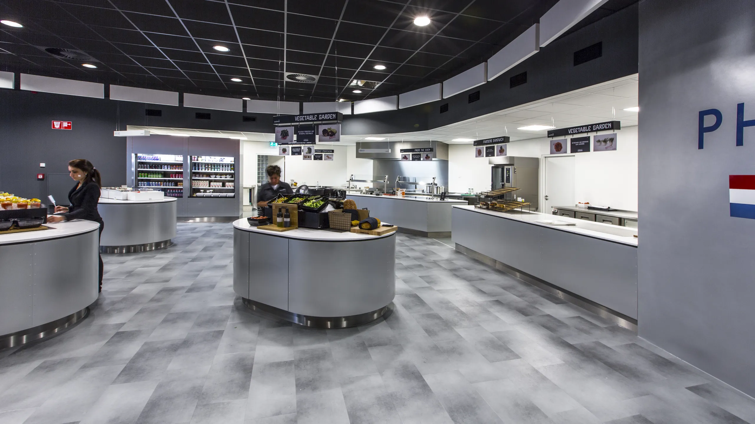 Self-service restaurant interior with food counters, circular serving stations, and a black acoustic grid ceiling above the dining area.