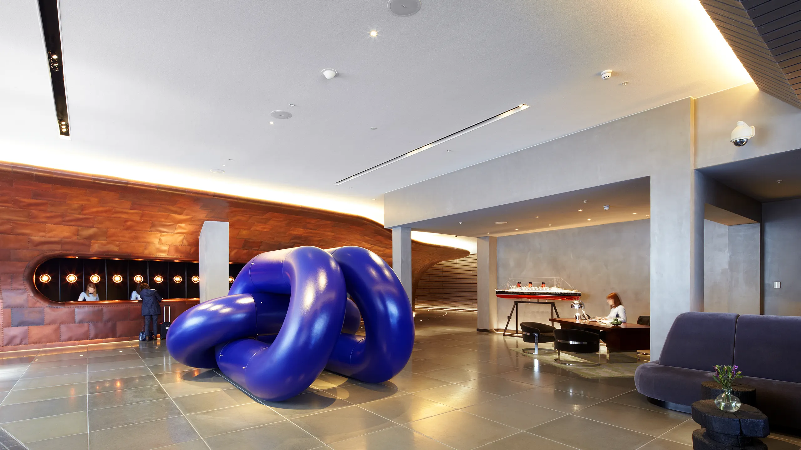 Hotel reception lobby with a smooth monolithic acoustic ceiling, curved copper-clad reception desk, and a large blue sculptural installation positioned in the centre of the space.