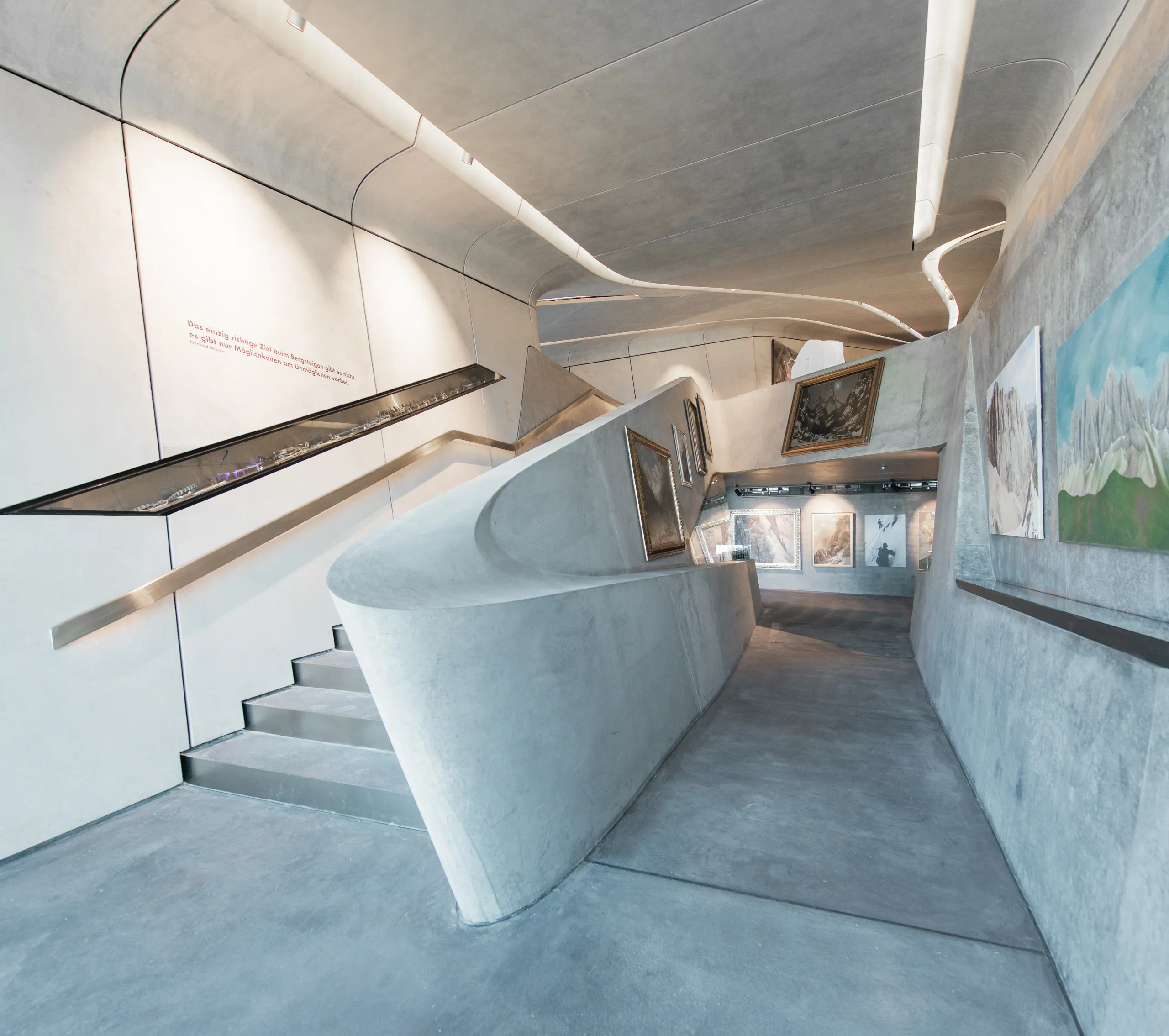 Concrete exhibition space with a large panoramic window overlooking a snowy mountain landscape, displaying sculptural pieces and interpretation panels inside a curved architectural interior.