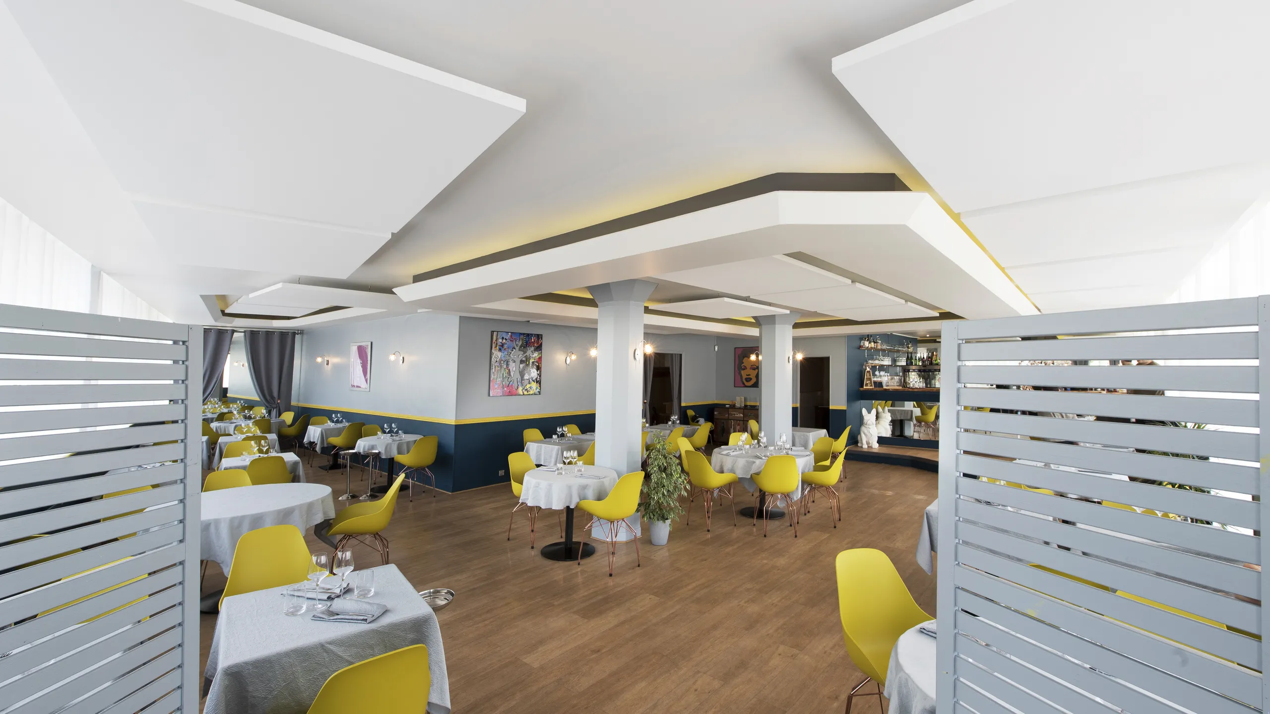Modern restaurant interior with yellow chairs, a floating raft ceiling, white tablecloths, wooden floor, contemporary decor, and soft lighting.