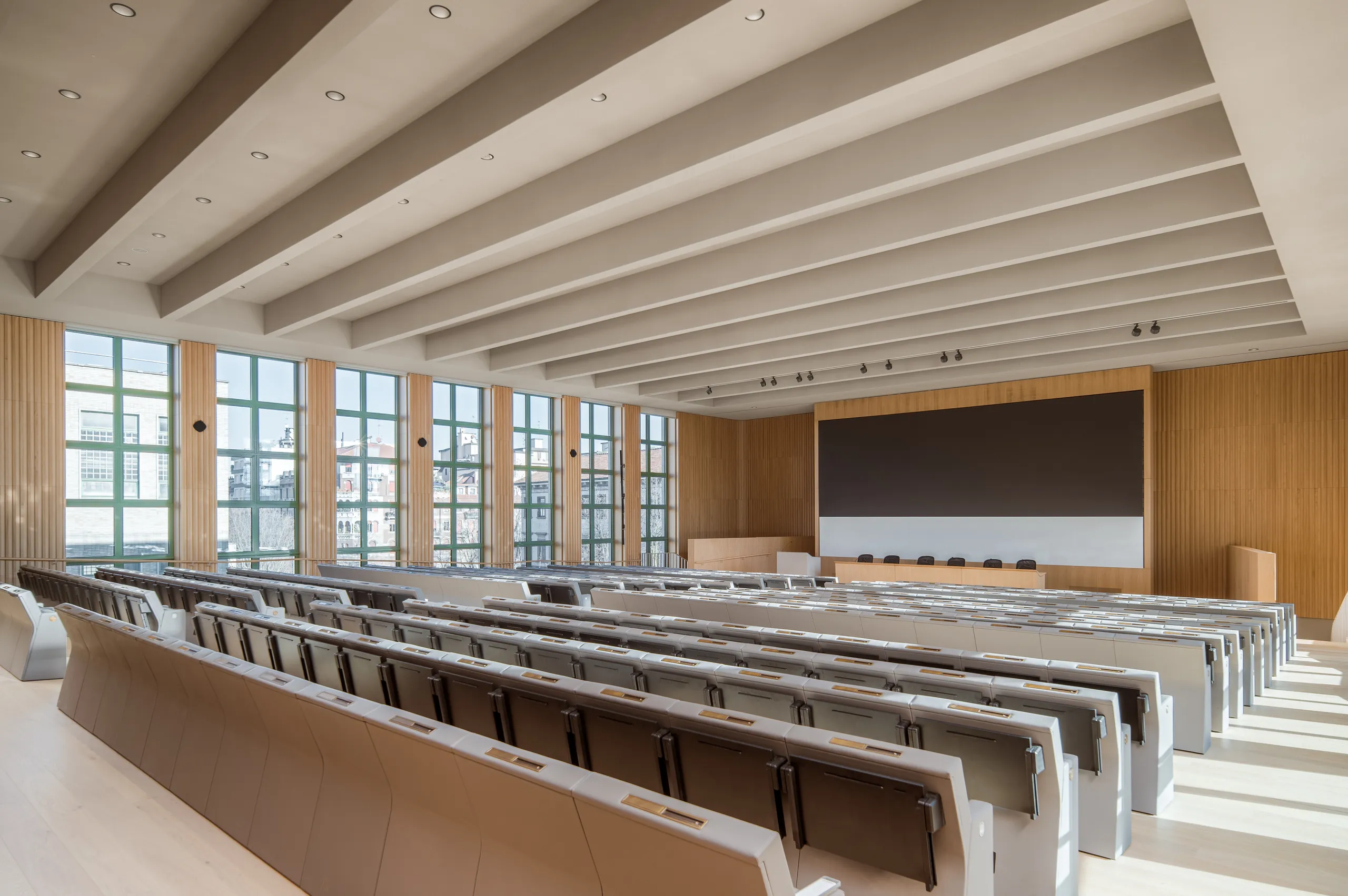 A spacious lecture hall with acoustic ceiling tiles placed between long exposed beams, above rows of tiered seating and a timber-lined presentation wall