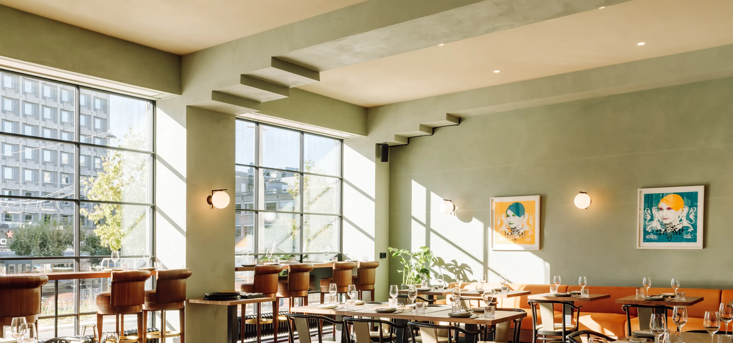 A bright, modern restaurant featuring a seamless monolithic acoustic ceiling in a soft neutral tone. The space includes large steel-framed windows, green plastered walls, warm wooden tables, bar seating, and framed artwork, with sunlight casting patterned shadows across the dining area.