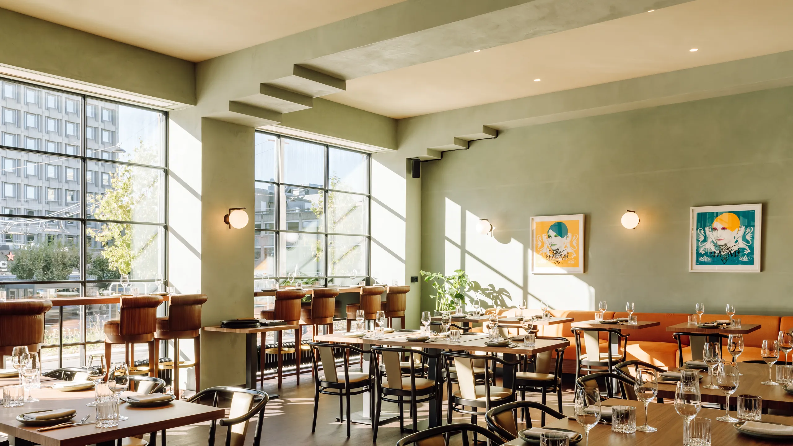 Modern restaurant interior with large windows, wooden tables, set with plates and glasses, soft lighting, and framed art on sage green walls.