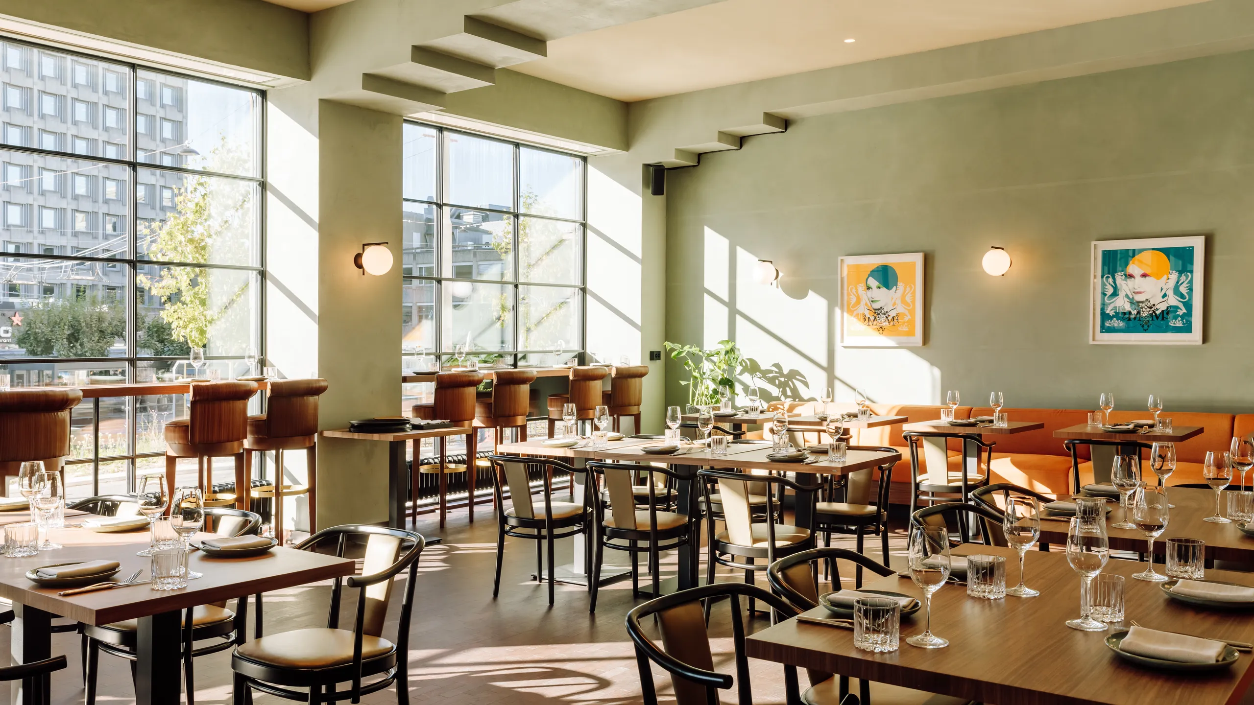 Sunlit restaurant interior with a smooth monolithic acoustic ceiling, green walls, large windows, wooden tables, and bar seating along the window, with artwork and plants throughout the space.