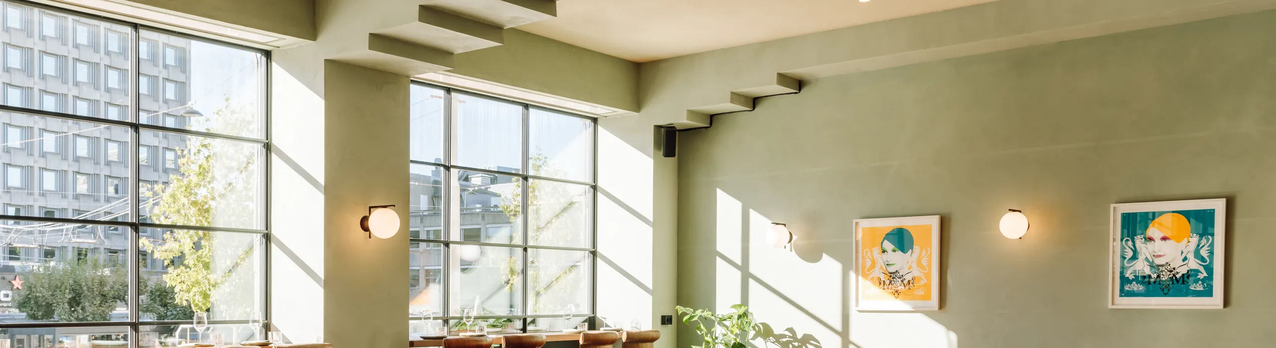 A bright, modern restaurant featuring a seamless monolithic acoustic ceiling in a soft neutral tone. The space includes large steel-framed windows, green plastered walls, warm wooden tables, bar seating, and framed artwork, with sunlight casting patterned shadows across the dining area.