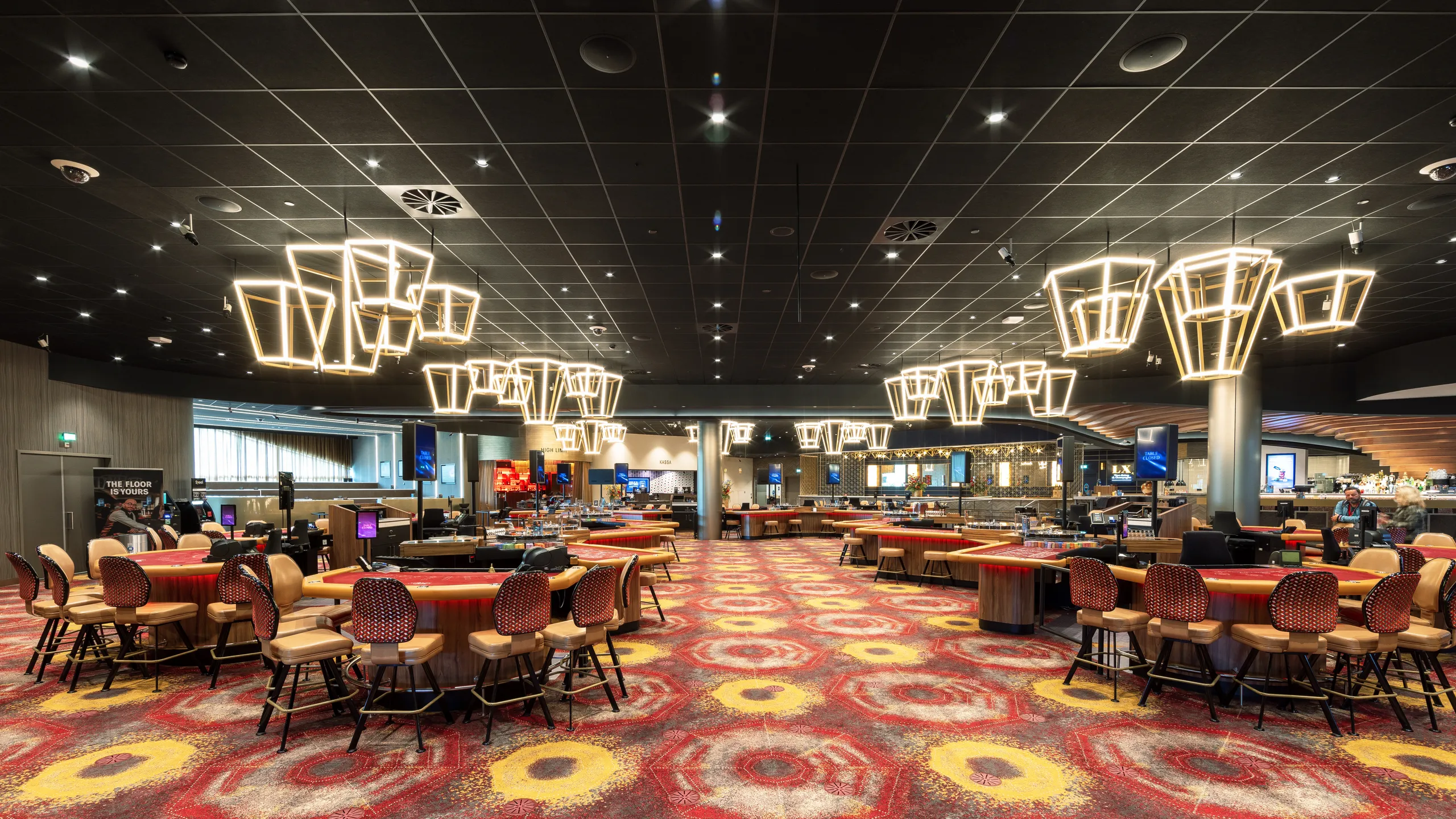 Spacious casino interior with a black suspended ceiling, modern geometric chandeliers, colorful patterned carpet, and rows of gaming tables and chairs.