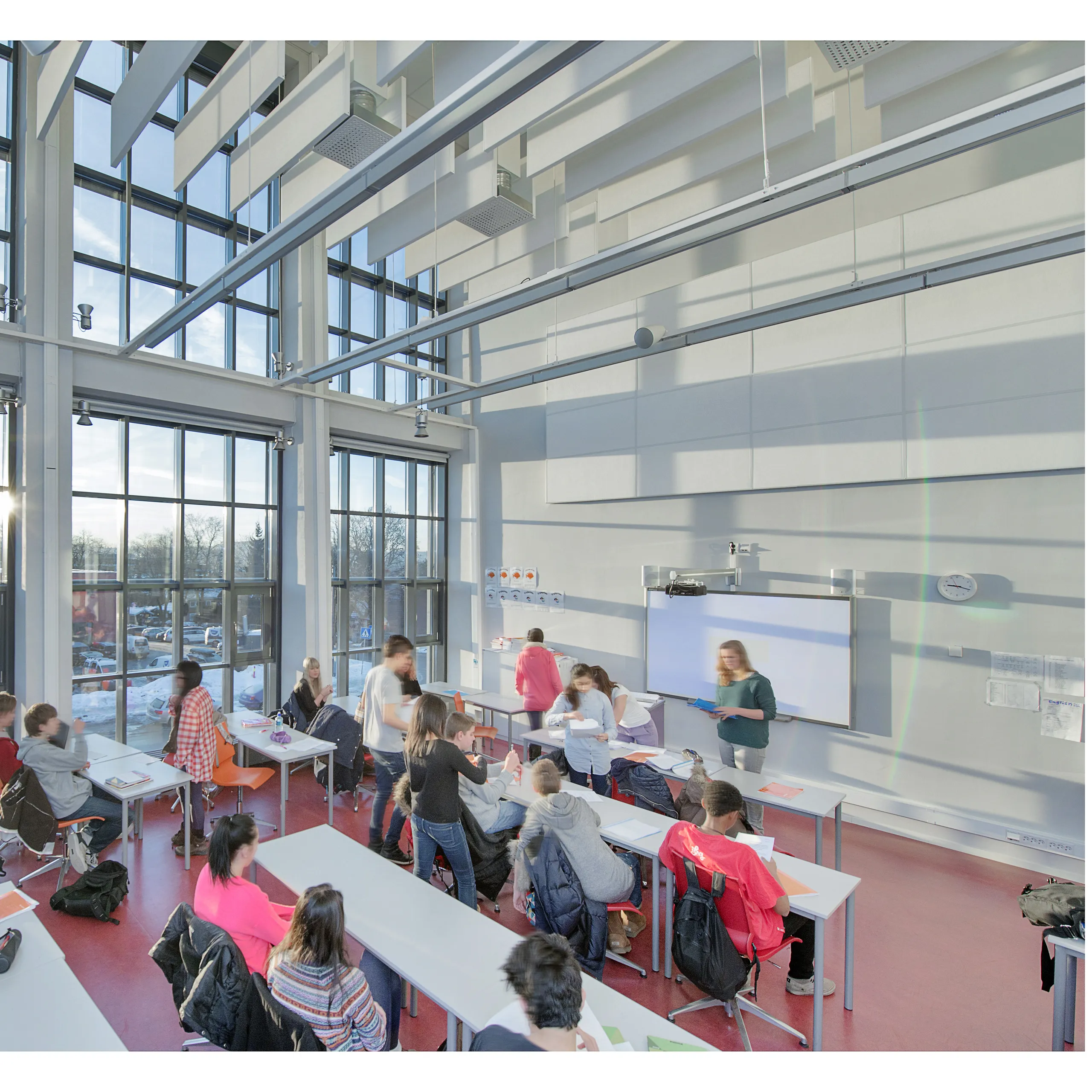 A large, light-filled classroom featuring suspended white acoustic baffles and exposed steel beams, with students working at tables below expansive floor-to-ceiling glazing.
