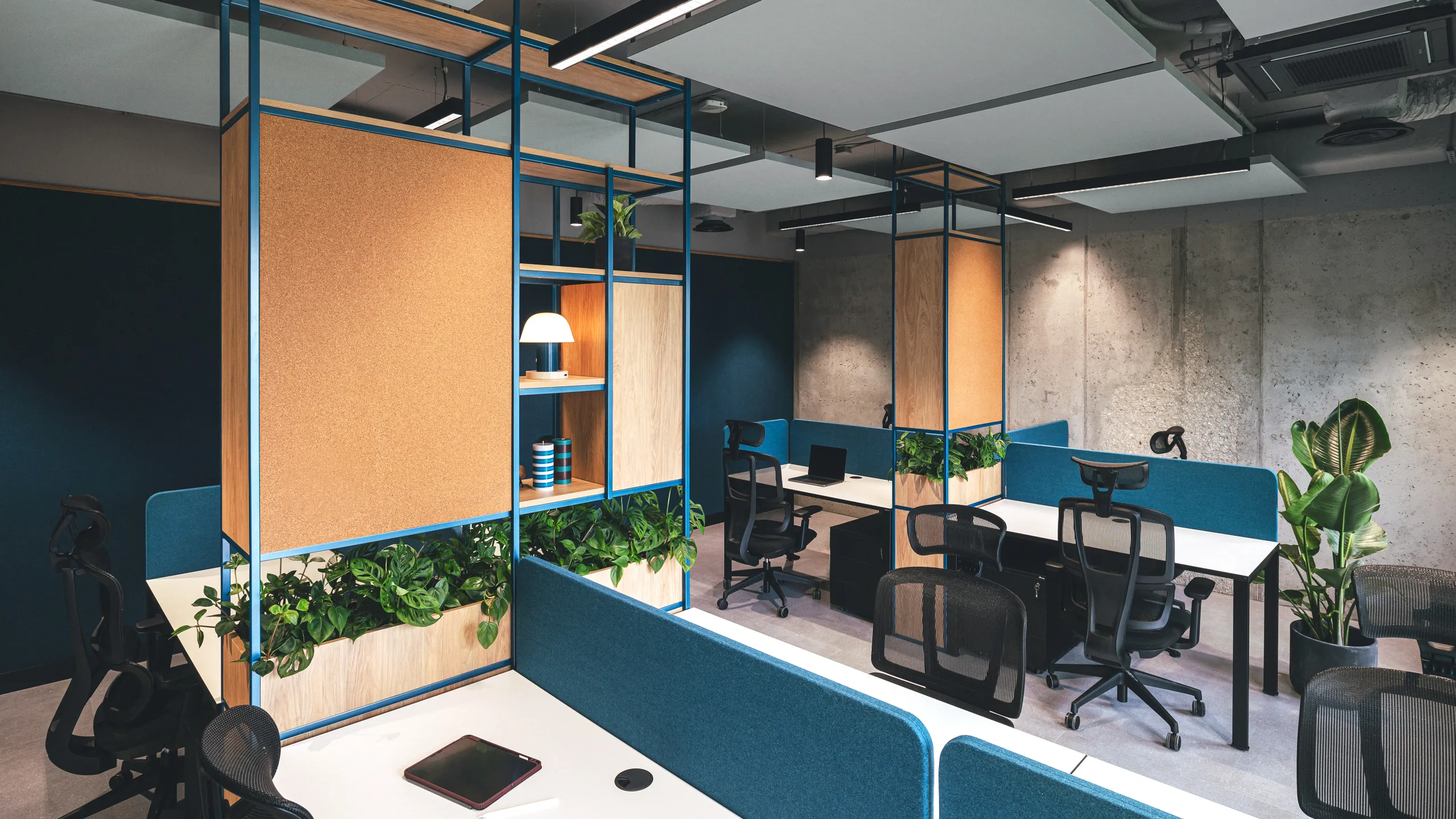 Modern office space with floating panels in the office ceiling, multiple workstations, blue partitions, plants, shelves, and overhead lighting in a stylish, open layout.