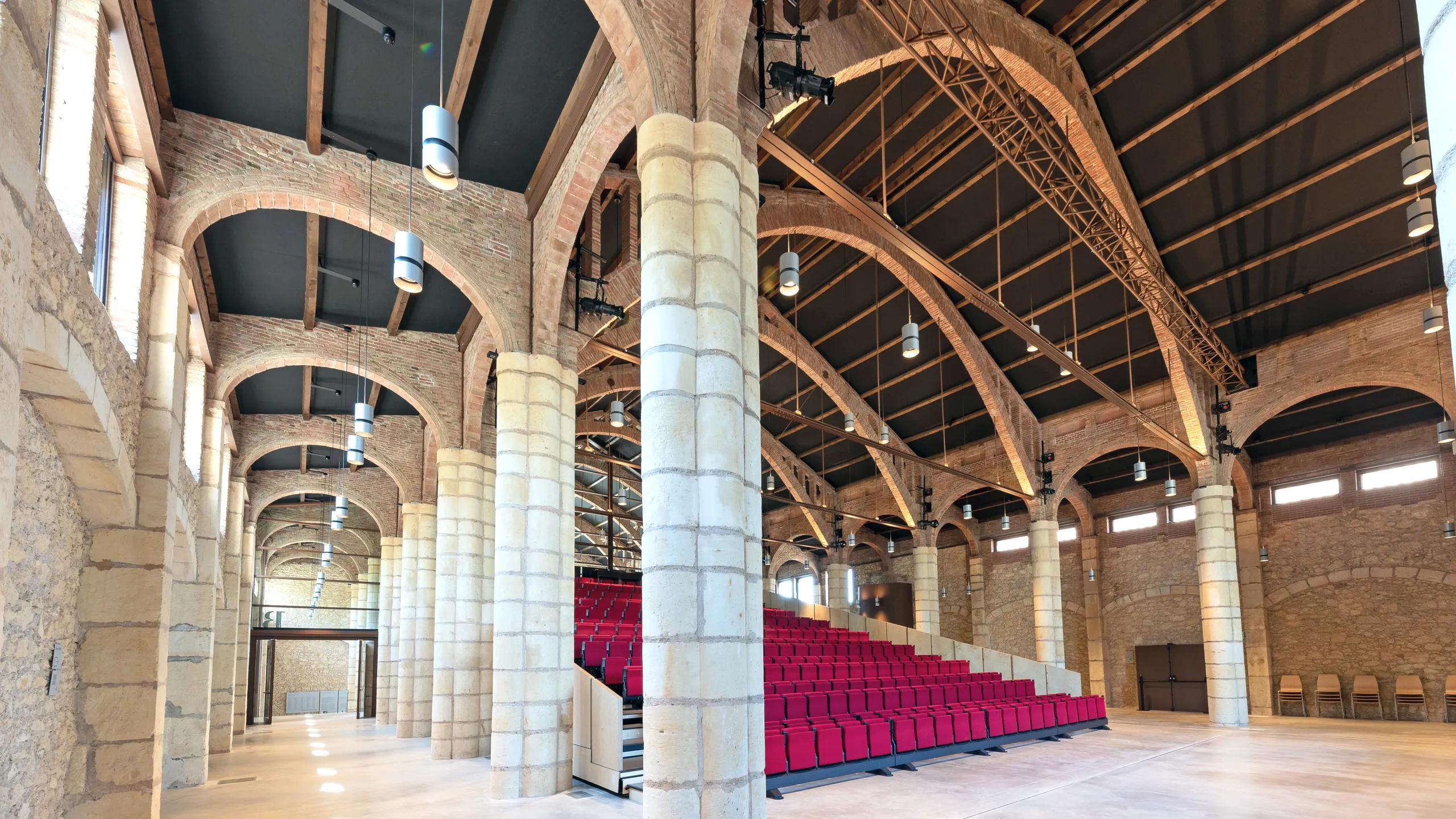 Auditorium with tiered red seating, a stage area, exposed brick arches, and black acoustic ceiling tiles fitted between the vaulted roof beams.