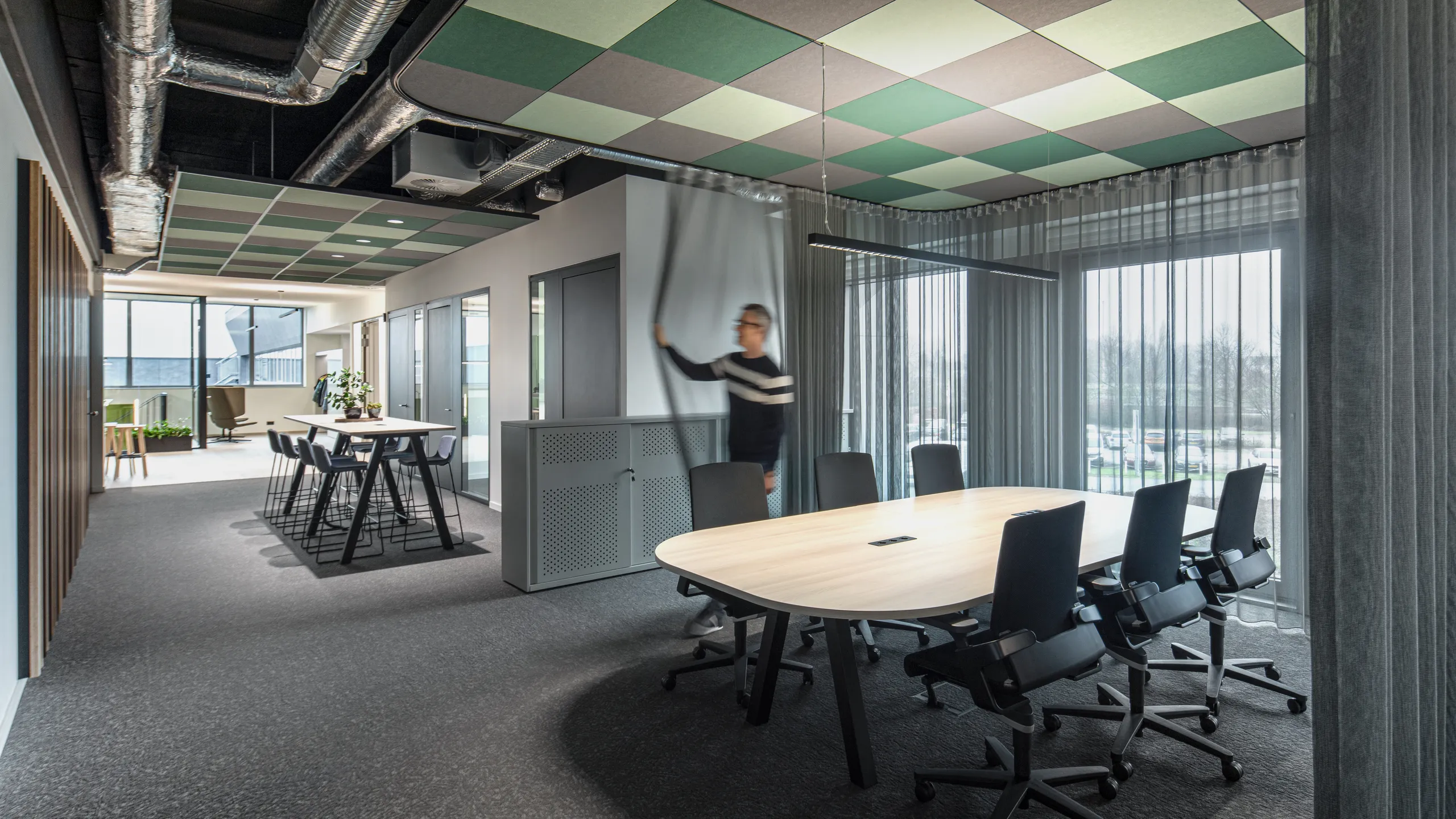 Modern office meeting area with a green and grey checkerboard acoustic ceiling, large meeting table, and collaborative workspace extending into the background.
