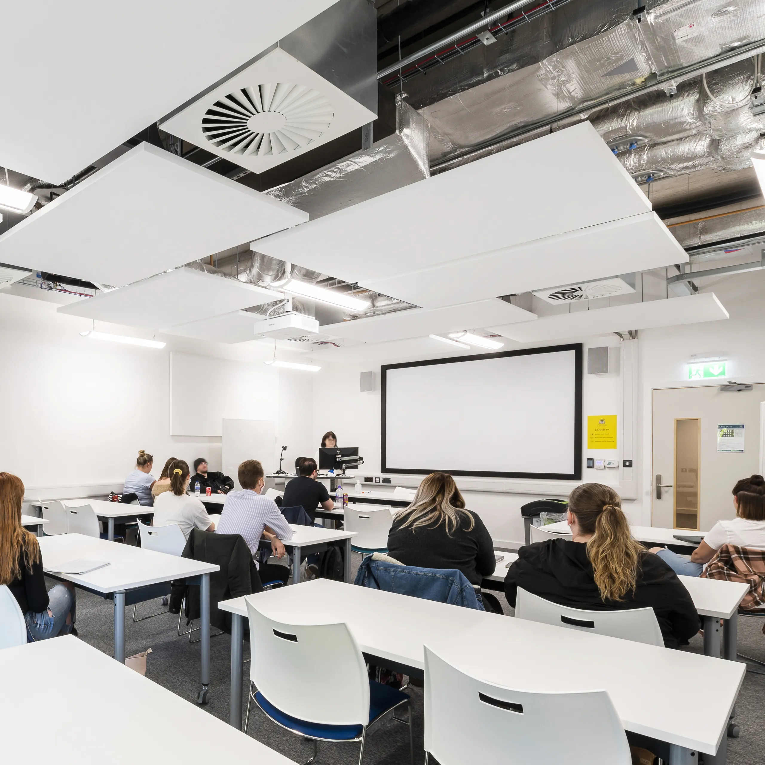 A modern classroom with students seated at white desks beneath suspended white acoustic ceiling rafts and exposed ductwork