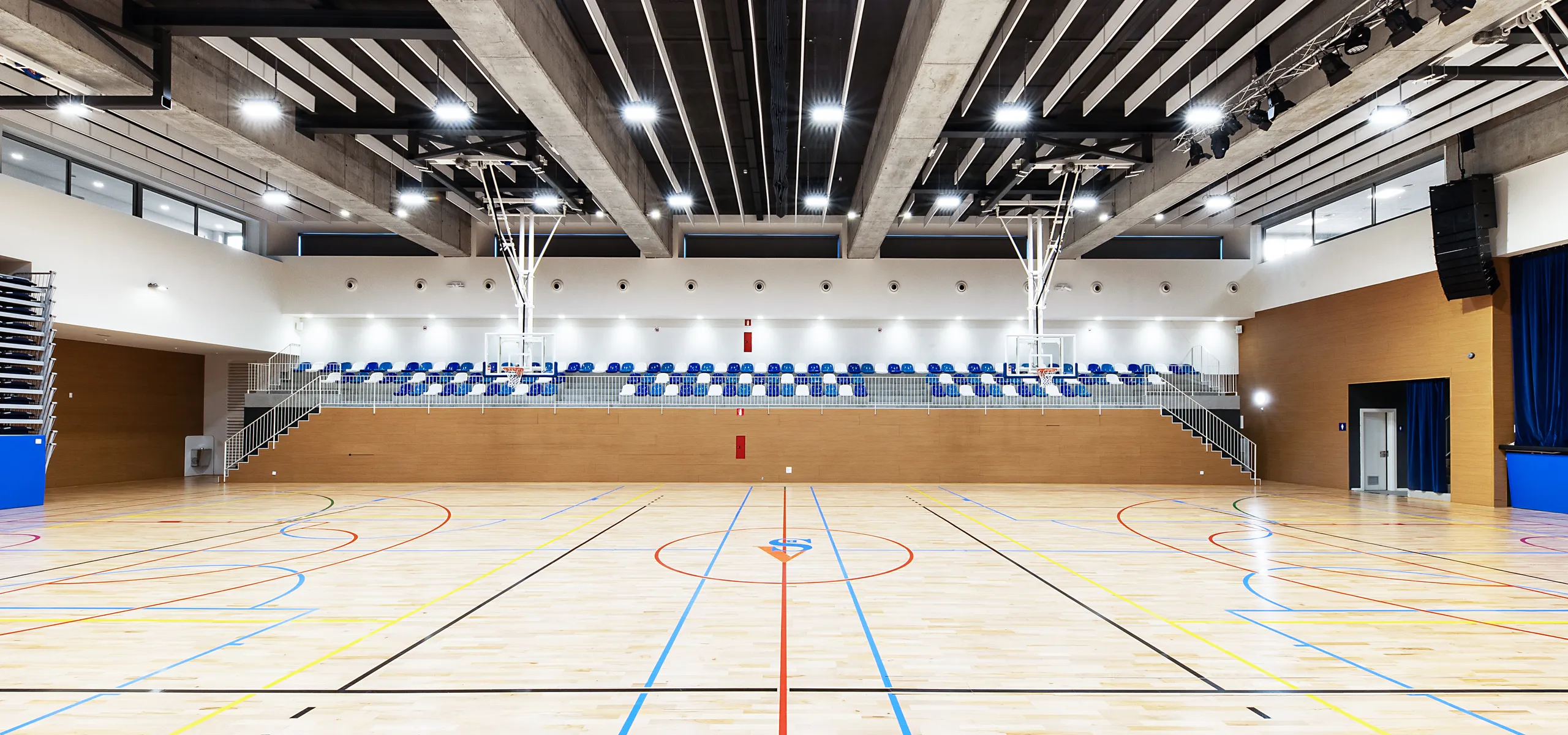 A spacious indoor sports hall featuring acoustic baffles installed between exposed concrete beams, with bright overhead lighting, blue seating stands, and a multi-line wooden court.