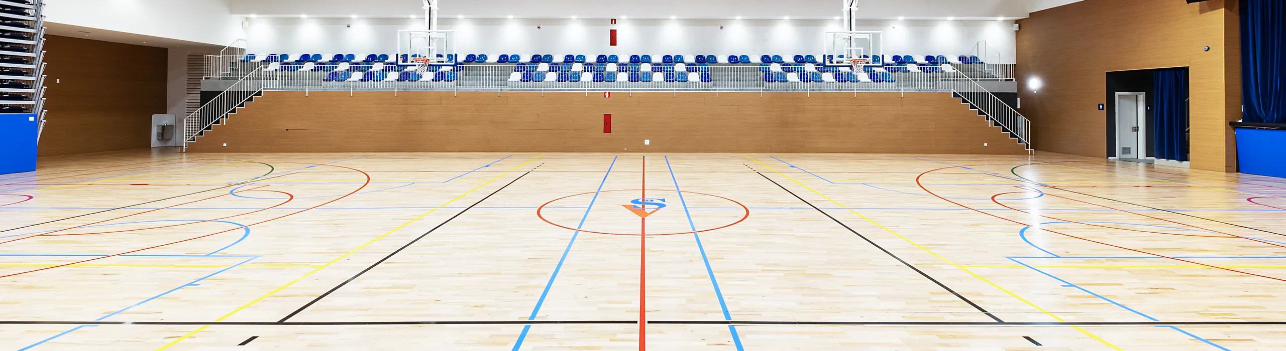 A spacious indoor sports hall featuring acoustic baffles installed between exposed concrete beams, with bright overhead lighting, blue seating stands, and a multi-line wooden court.