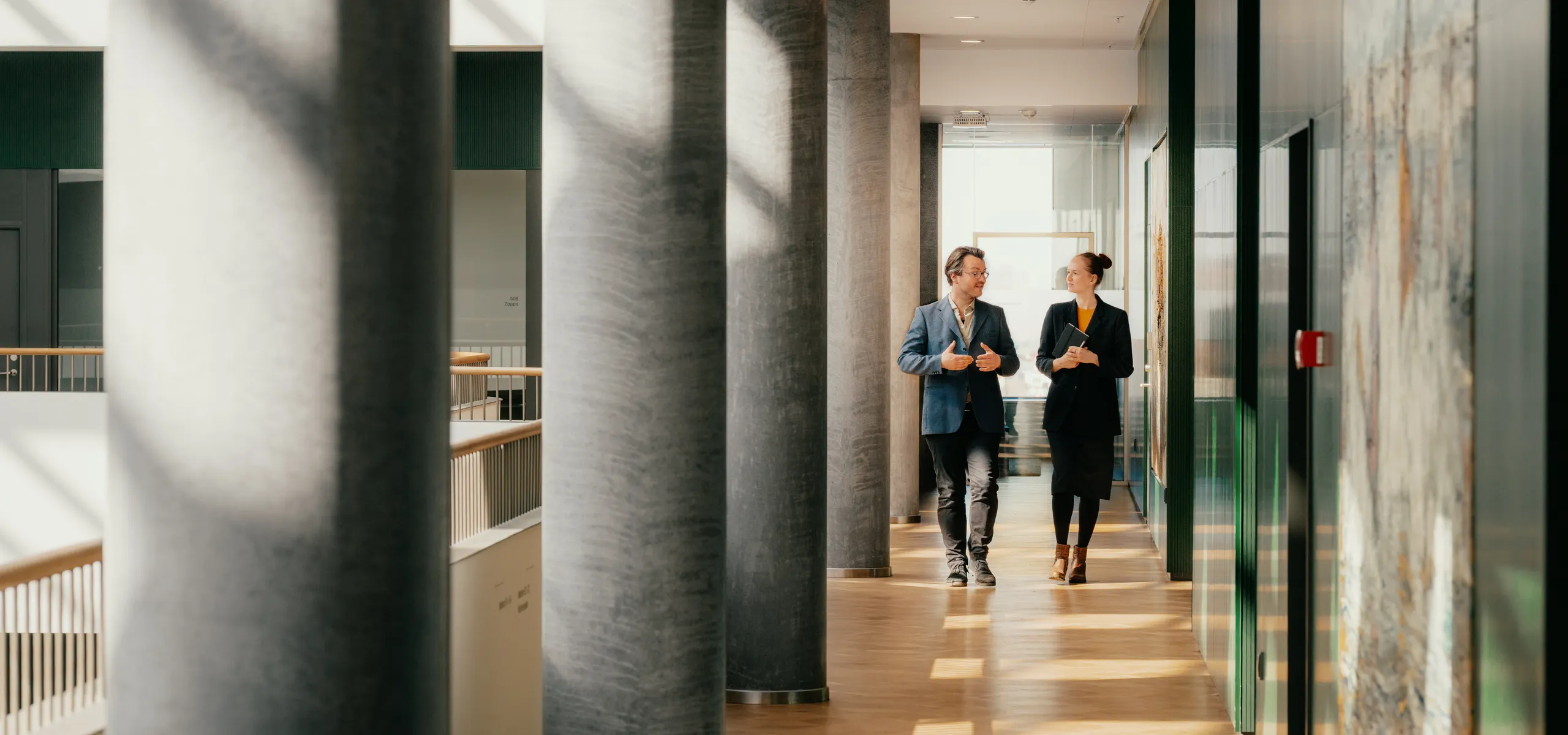 Two people walking and talking in a bright hallway with white acoustic modular ceilings, large columns and artwork, illuminated by sunlight streaming through windows.